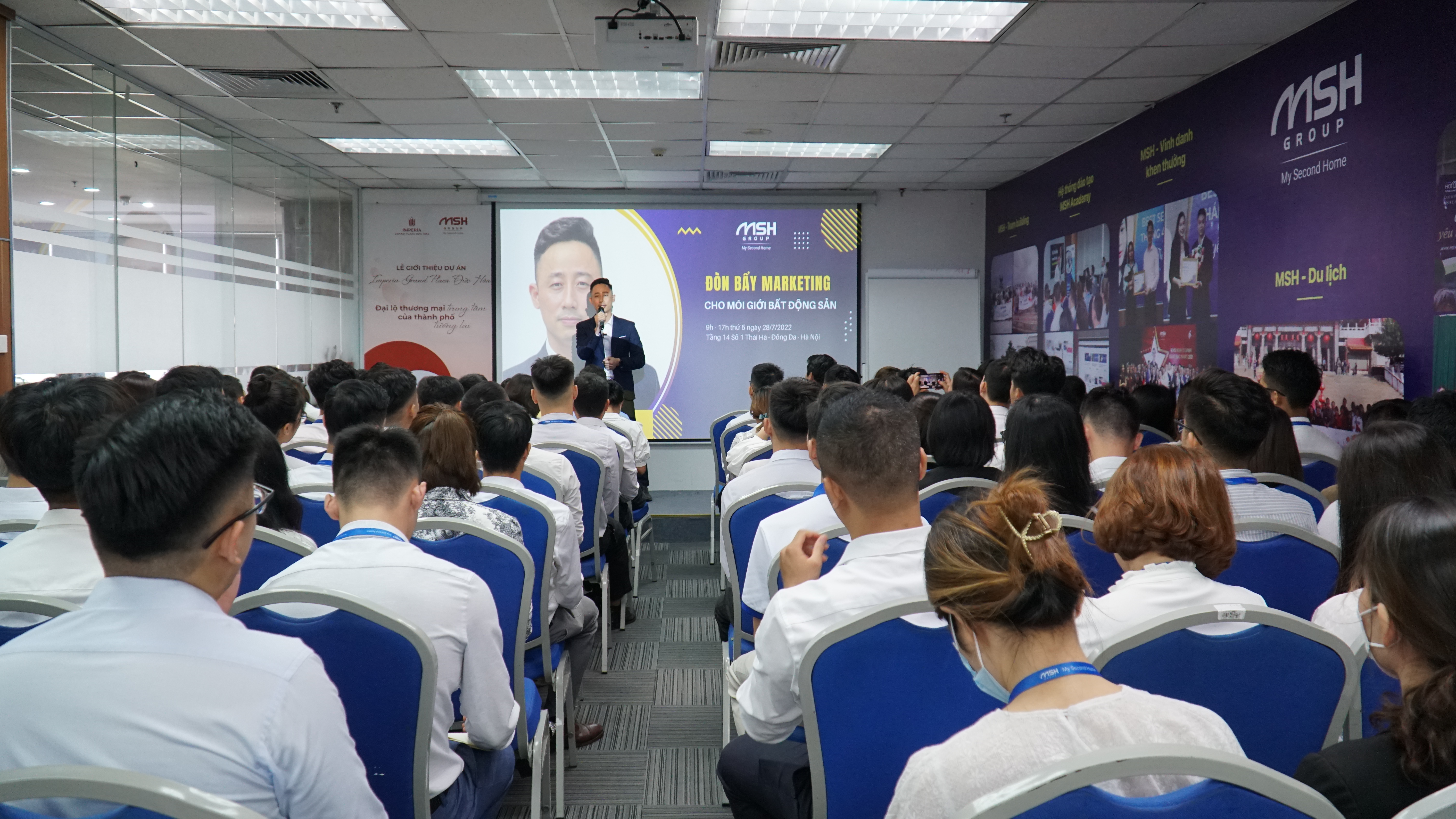 Toàn cảnh một phòng đào tạo đông kín, khoảng sáu mươi môi giới mặc đồng phục sơ mi trắng ngồi trên ghế xanh cùng hướng về phía màn chiếu và người giảng ở đầu phòng