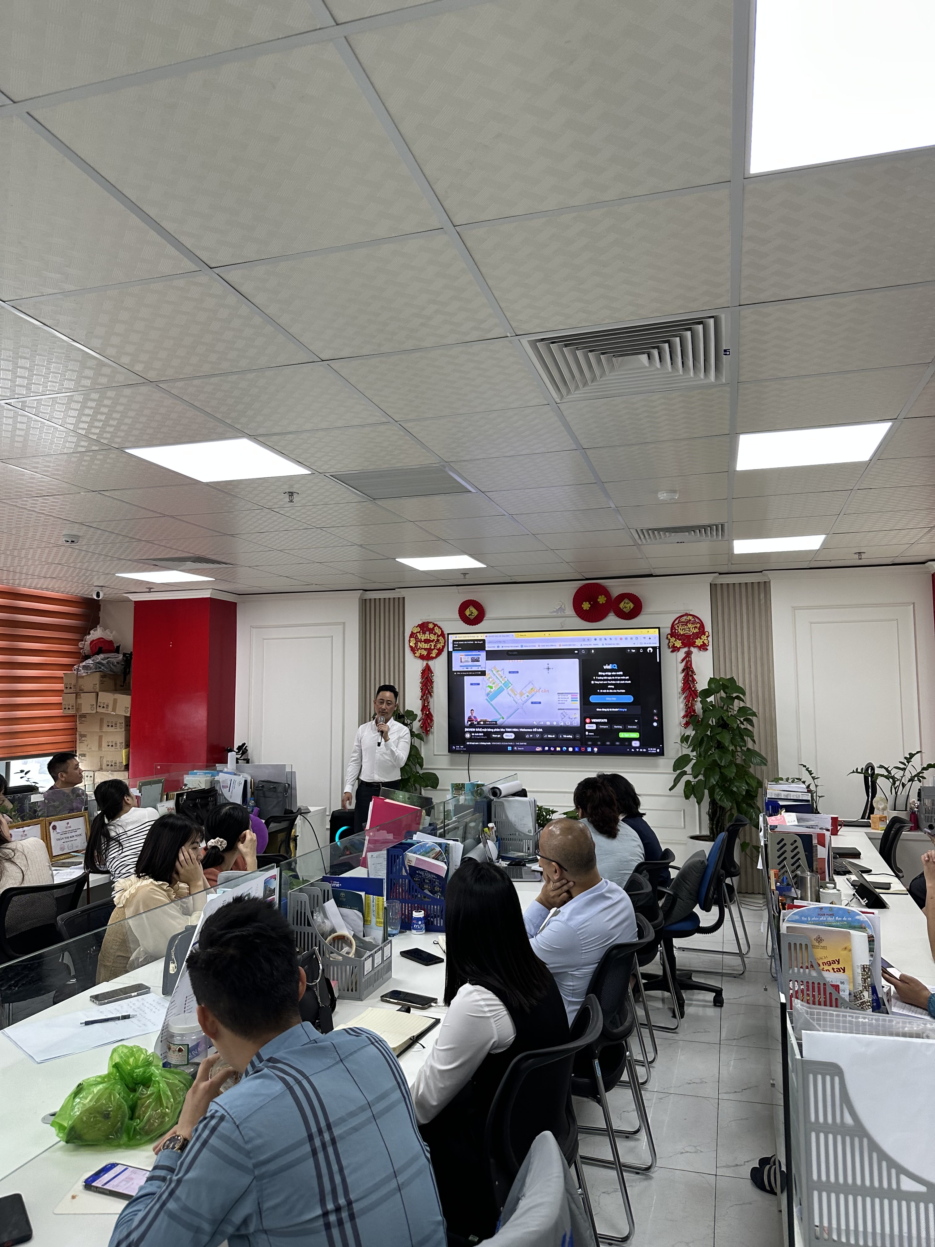 Một văn phòng mở trần đèn panel, Đăng Dương mặc áo trắng đứng nói trước màn hình treo tường đang chiếu bản đồ dự án, khoảng mười lăm người ngồi tại bàn làm việc quay về phía màn hình