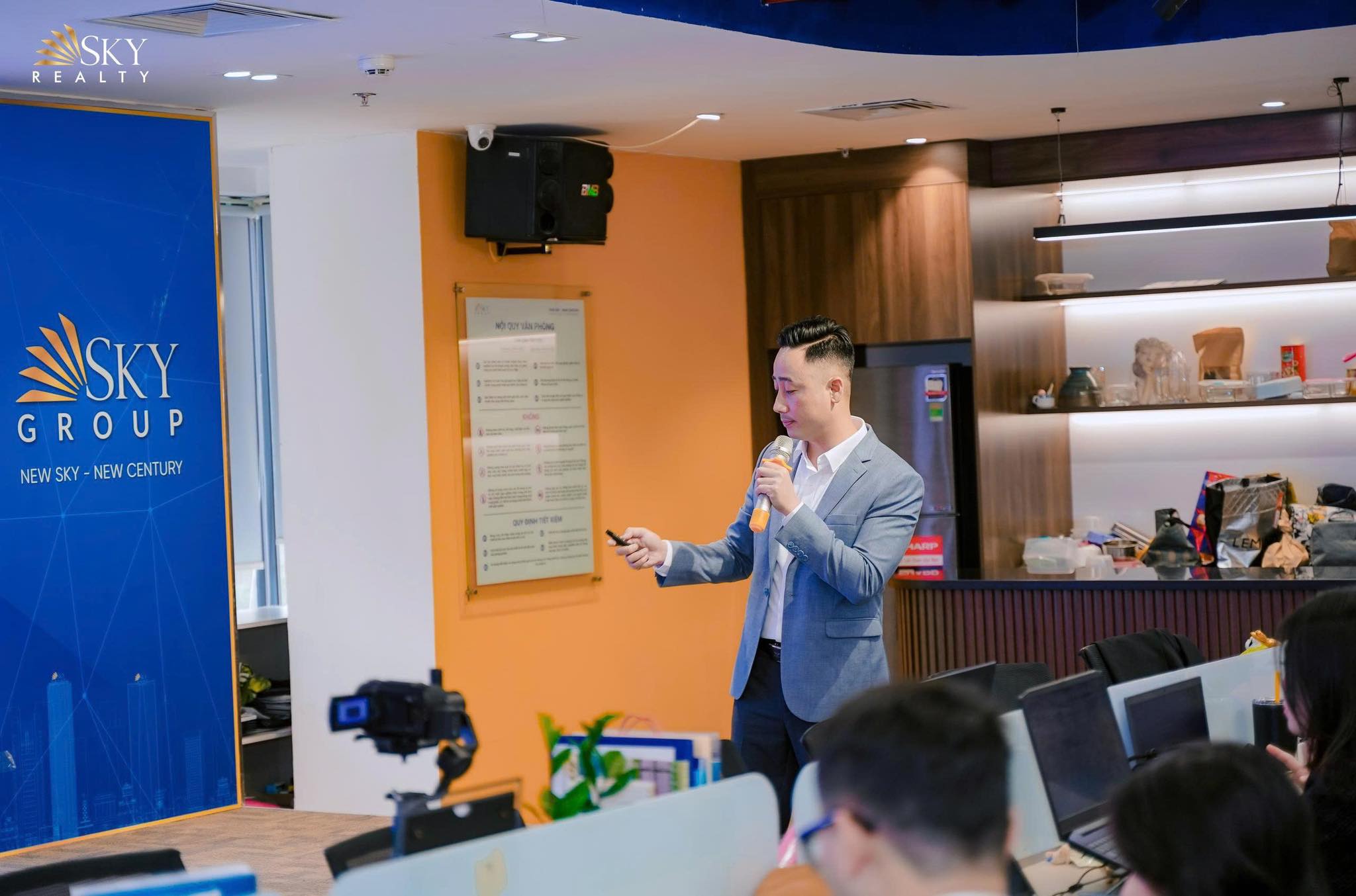 Đăng Dương mặc vest xám cầm micro nói trước đội ngũ đang ngồi ở bàn máy tính trong văn phòng Sky Group, một máy quay đặt sẵn trên chân máy phía trước