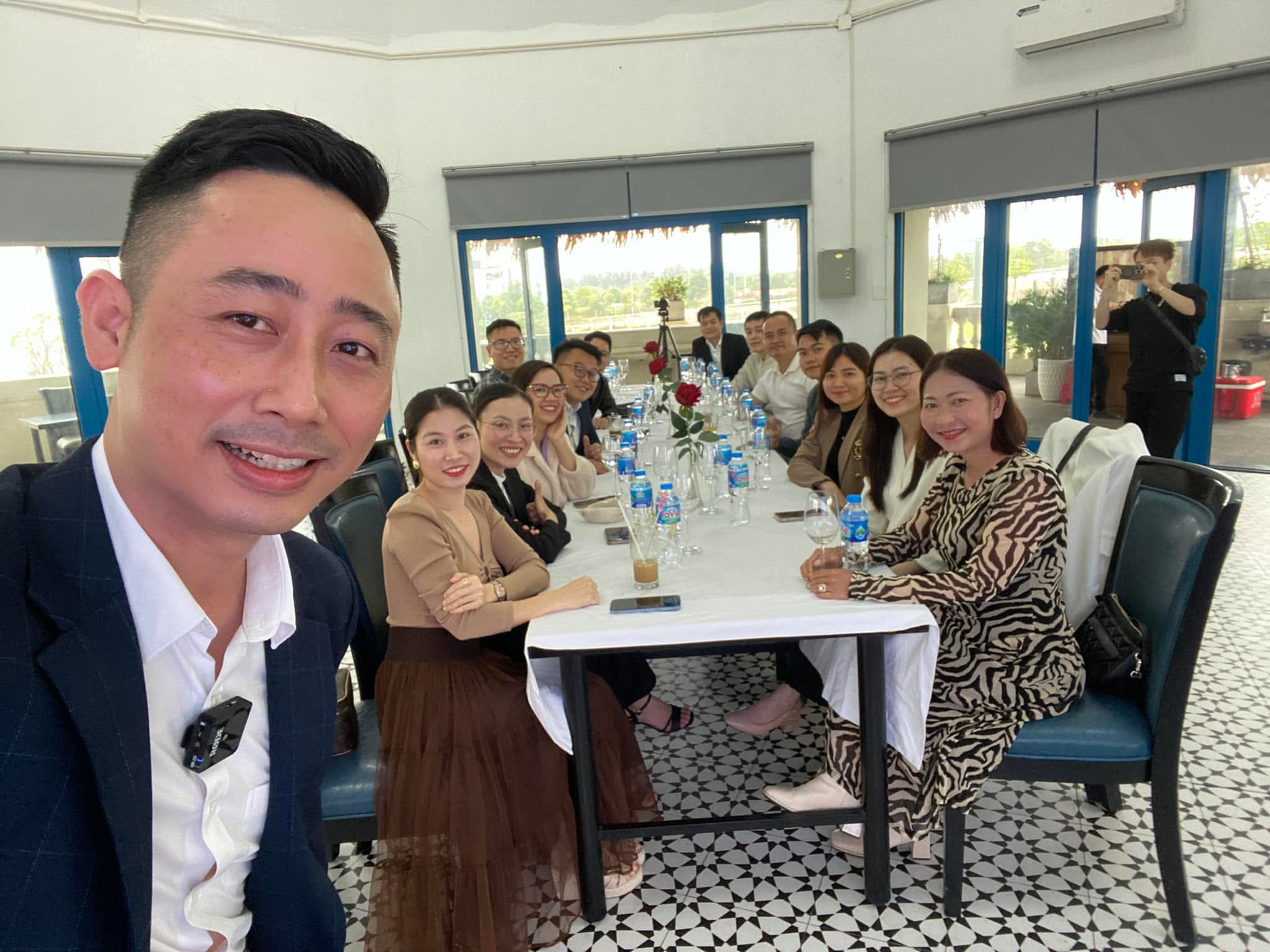 Đăng Dương mặc vest xanh than gắn mic cài áo chụp ảnh cùng hơn mười người ngồi hai bên một bàn dài phủ khăn trắng trong phòng ăn trên tầng cao