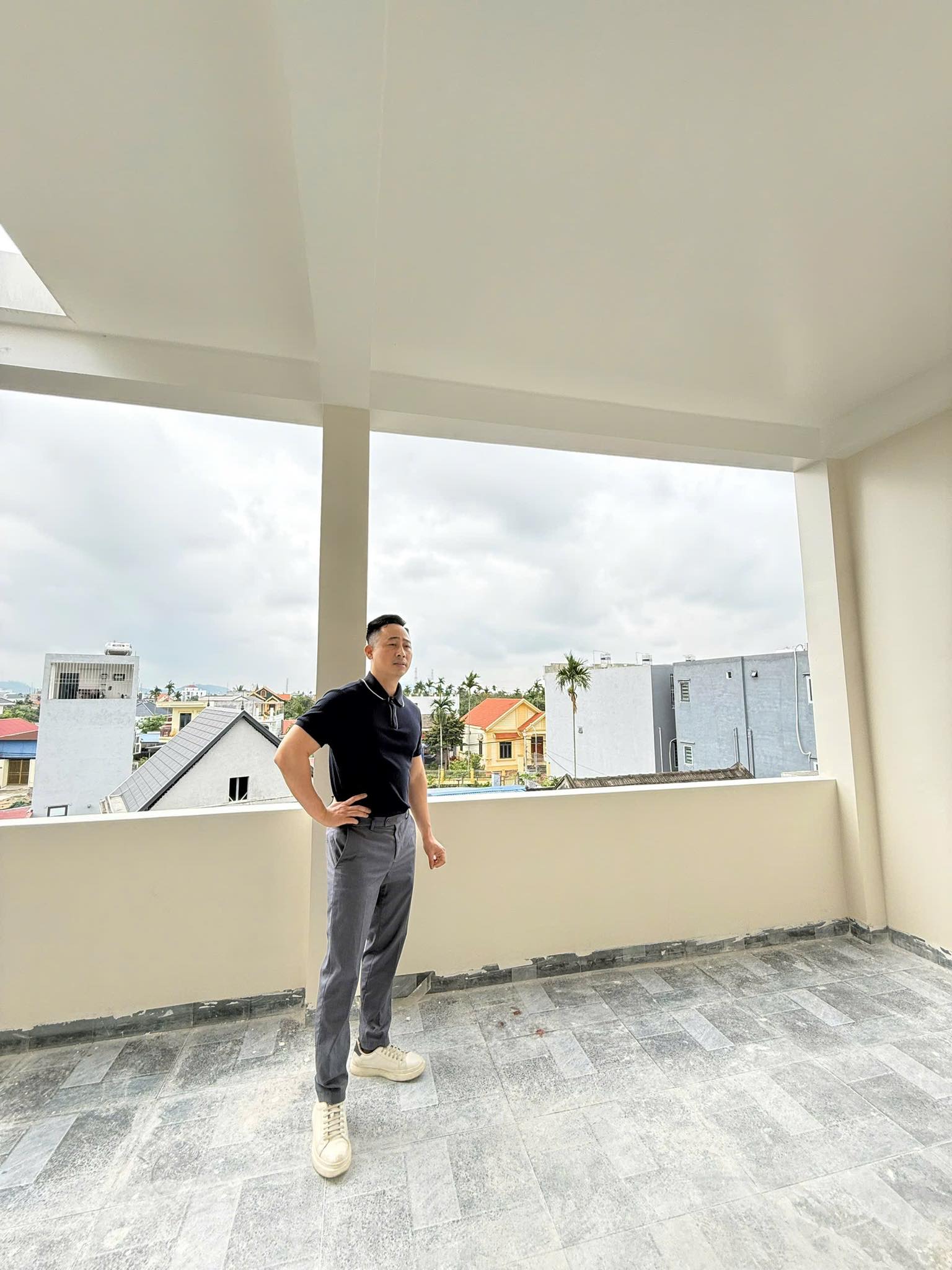 Đăng Dương mặc áo polo đen đứng chống một tay lên hông giữa căn phòng vừa lát nền của một ngôi nhà đang hoàn thiện, phía sau là ô cửa lớn nhìn ra mái nhà trong khu dân cư