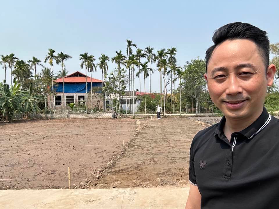 Đăng Dương mặc áo polo đen đứng cận cảnh trước một lô đất đã san phẳng, phía sau là hàng cau và một căn nhà đang xây phủ bạt xanh