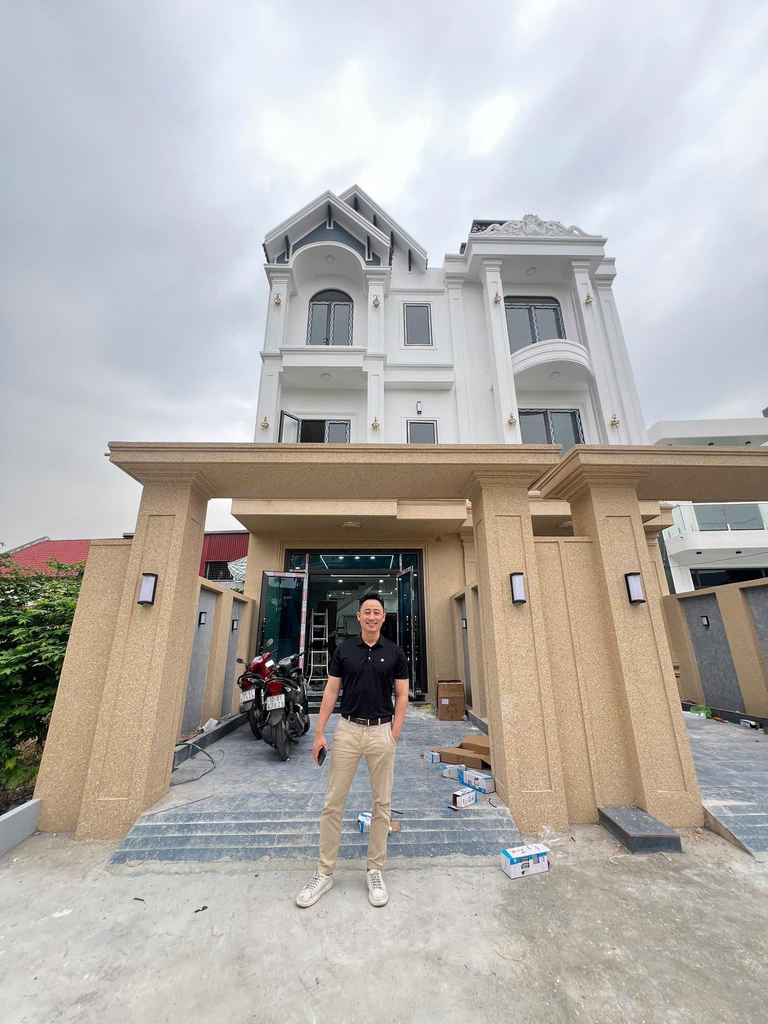 Đăng Dương mặc áo polo đen đứng trước một căn nhà ba tầng vừa hoàn thiện, thợ đang lắp cửa kính bên trong
