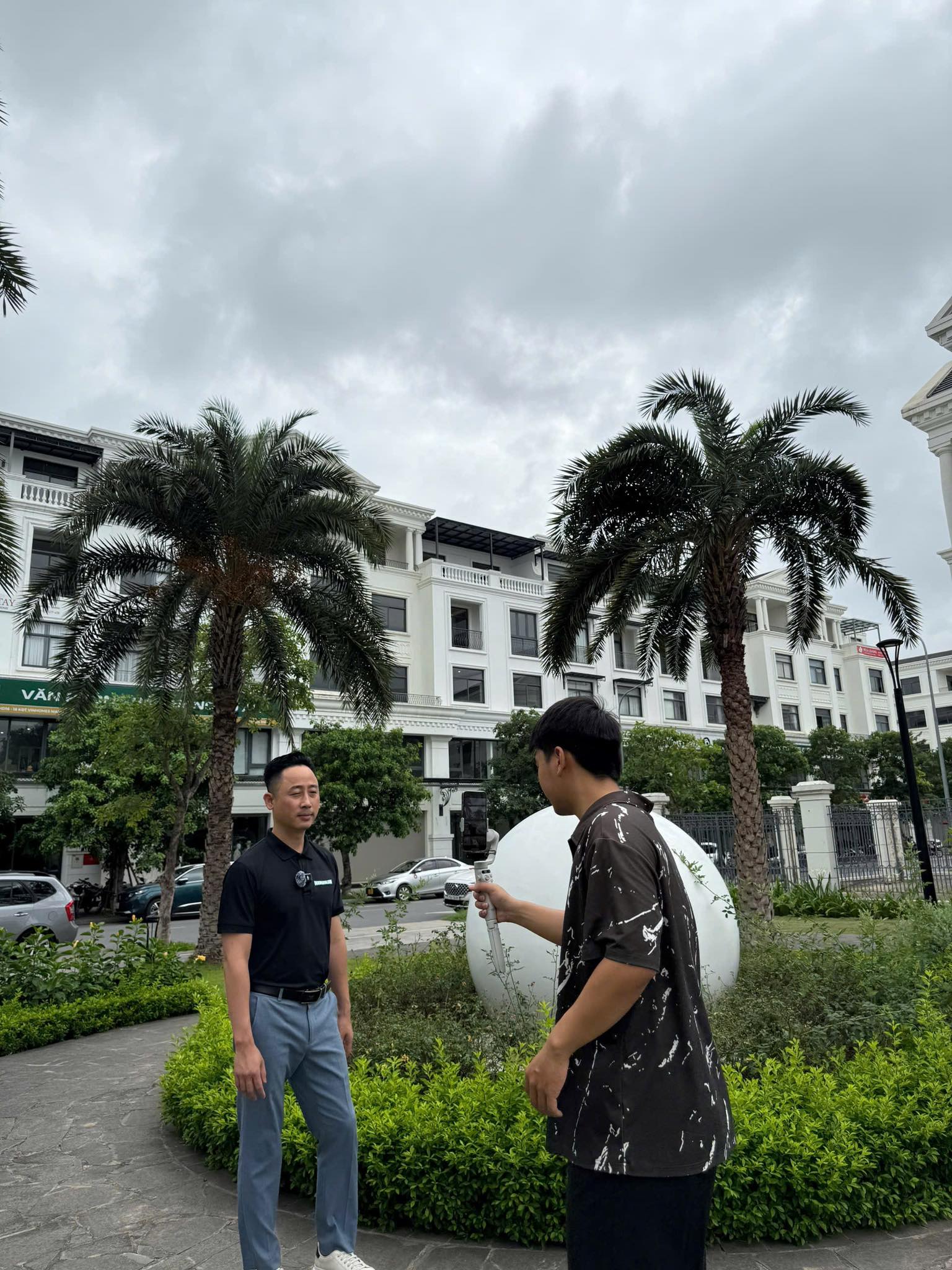 Đăng Dương mặc áo polo đen đứng trước máy quay do một bạn trẻ cầm gimbal, tại quảng trường một khu đô thị có hàng cọ
