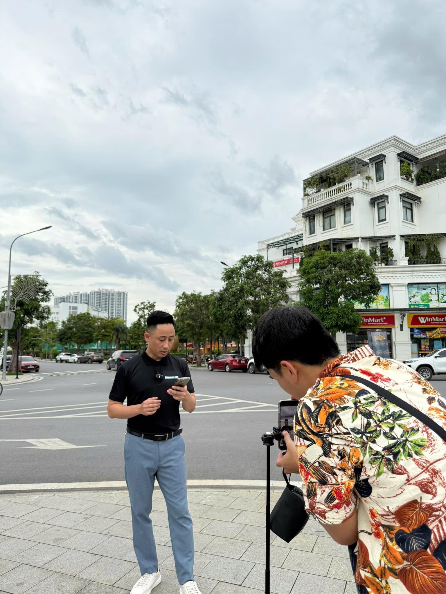 Đăng Dương mặc áo polo đen cầm điện thoại đứng bên đường tại một ngã tư khu đô thị, phía trước là một người mặc áo hoa cúi xuống quay bằng máy gắn gimbal, phía sau là dãy shophouse trắng