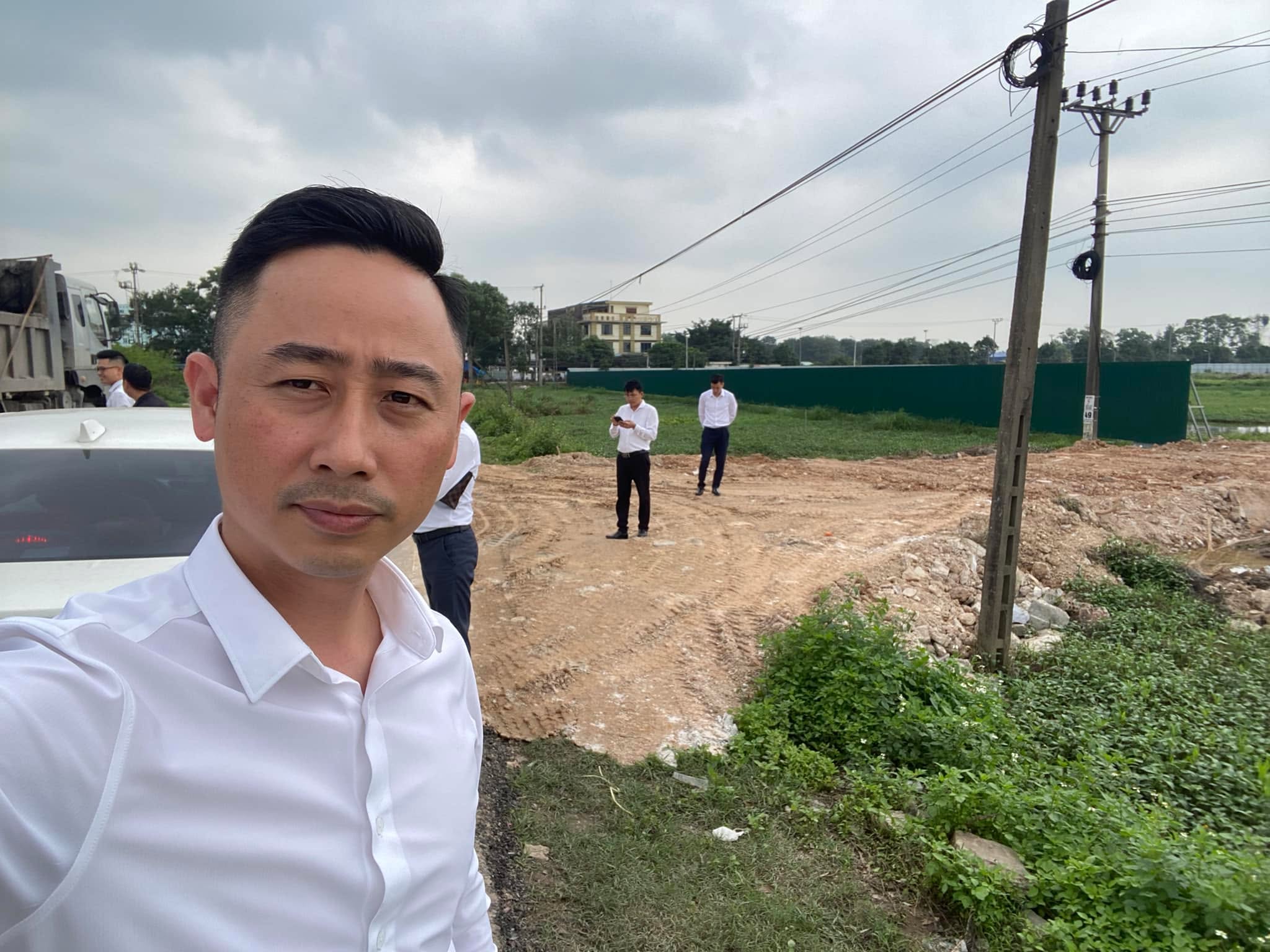 Đăng Dương mặc sơ mi trắng tự chụp ảnh trước một khu đất đang san lấp, phía xa có hai người mặc sơ mi đứng trên nền đất, bên trái là xe tải và hàng rào tôn xanh