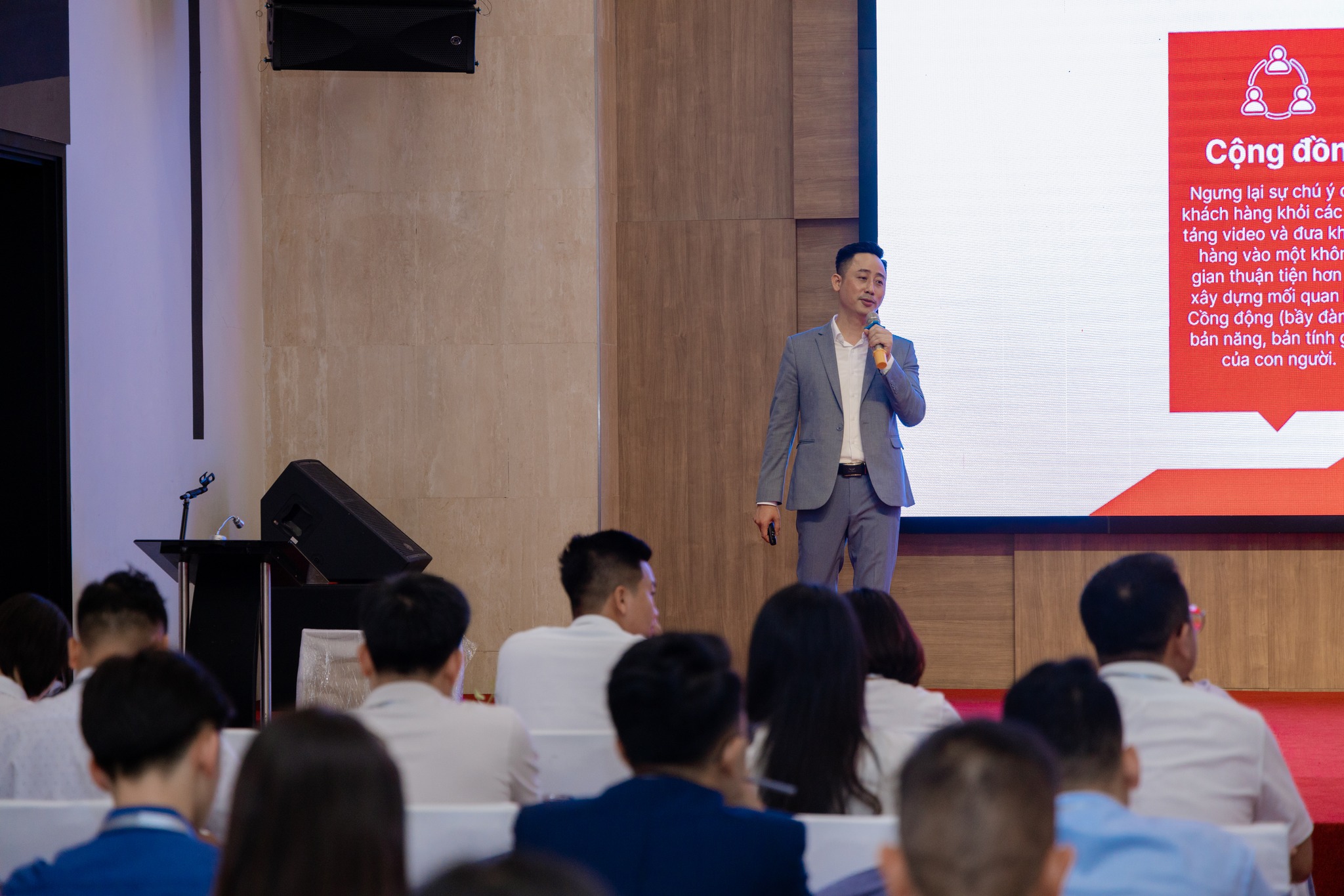 Đăng Dương mặc vest xám cầm micro đứng nói trên sân khấu hội trường, bên phải là màn chiếu đang hiện một slide nền đỏ, phía dưới là khán giả ngồi kín ghế