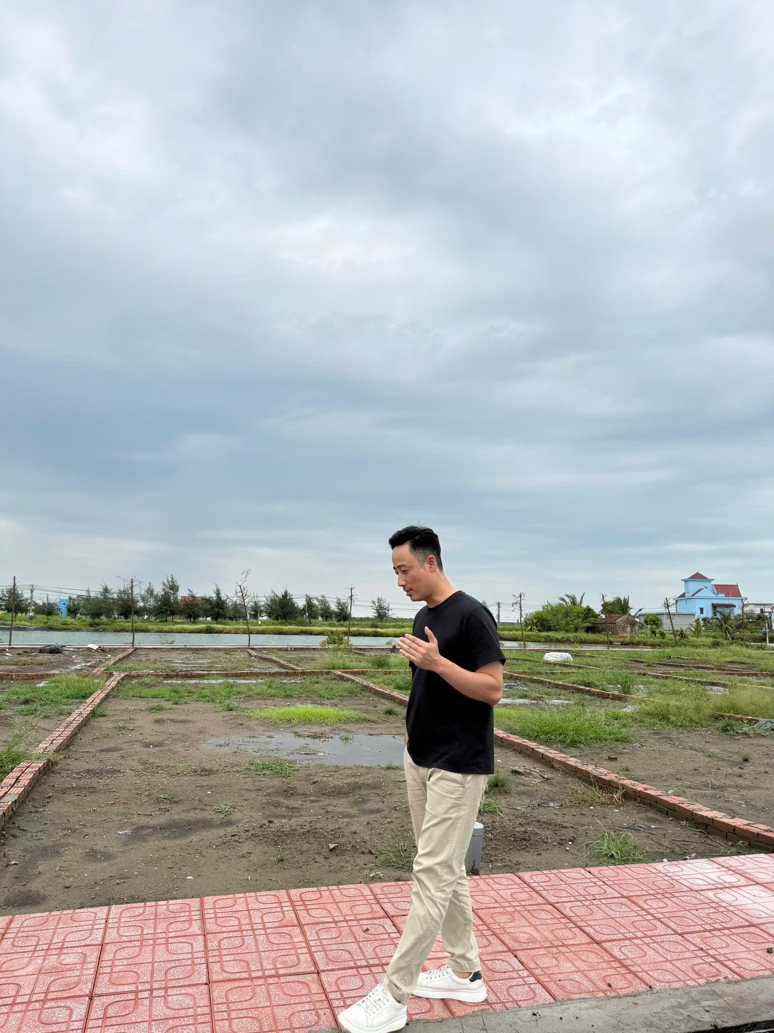 Đăng Dương mặc áo phông đen đi bộ trên lối gạch cạnh khu đất phân lô ven sông