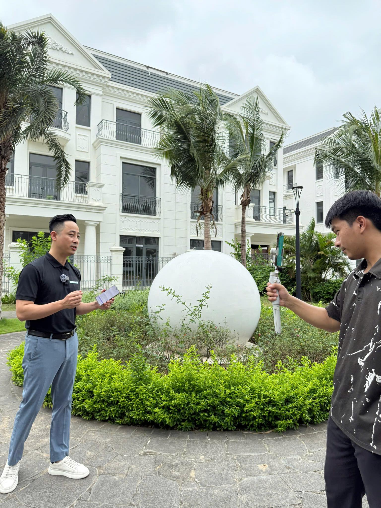 Đăng Dương mặc áo polo đen cầm điện thoại, đứng nói trước máy quay gimbal tại quảng trường một dự án biệt thự