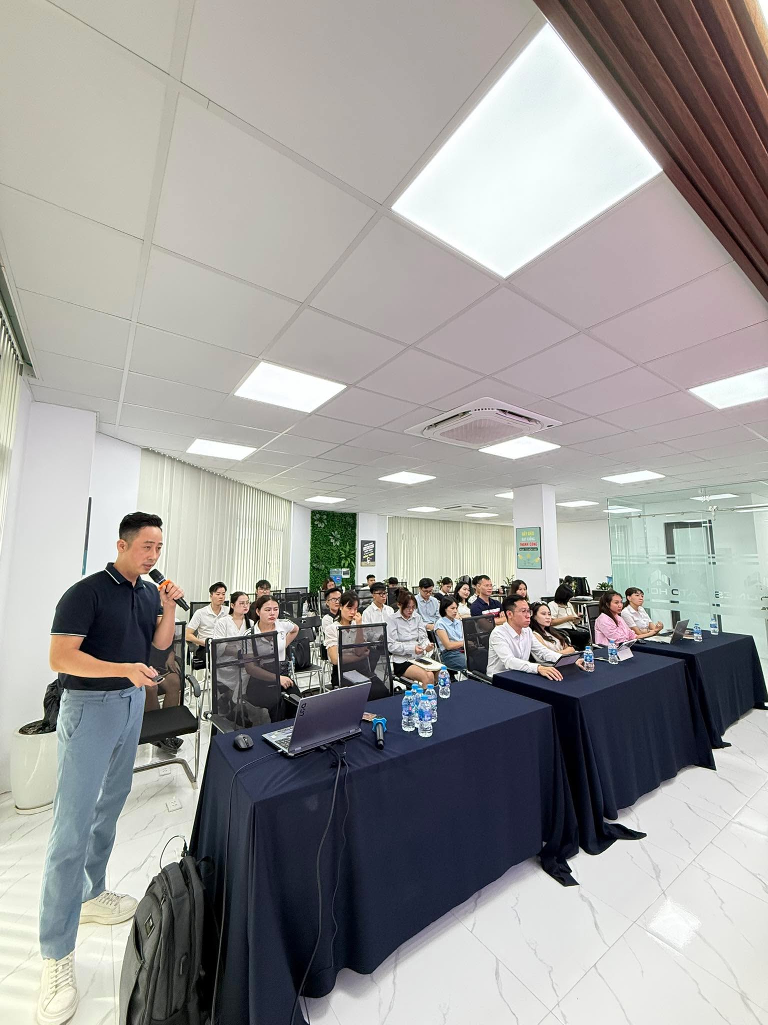 Đăng Dương cầm micro đứng giảng trước một lớp học đông người trong văn phòng