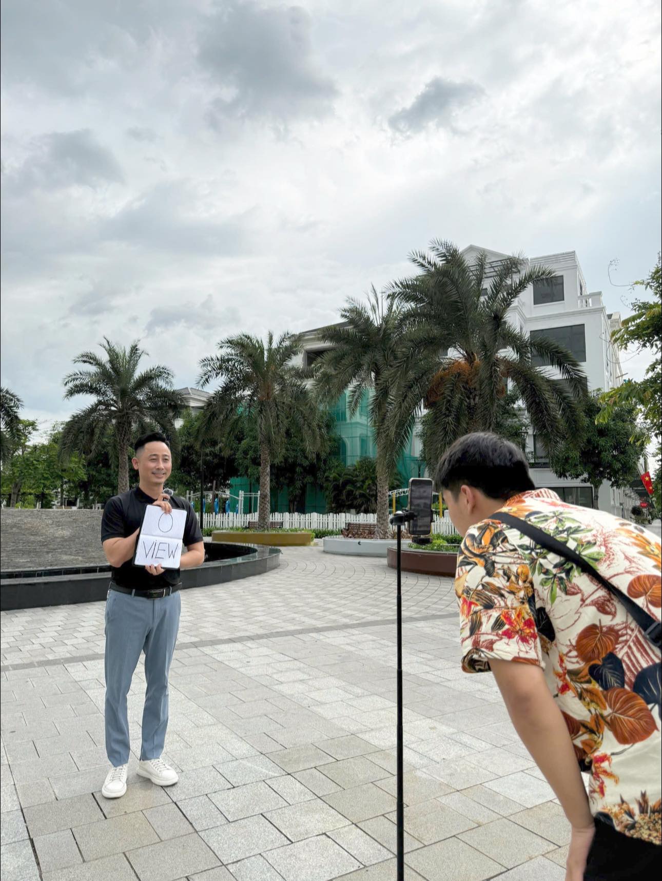 Đăng Dương mặc áo polo đen cầm tấm bảng viết chữ 0 VIEW đứng quay video trước điện thoại đặt trên chân máy giữa quảng trường khu đô thị, một người mặc áo hoa đang cúi xuống canh khung hình