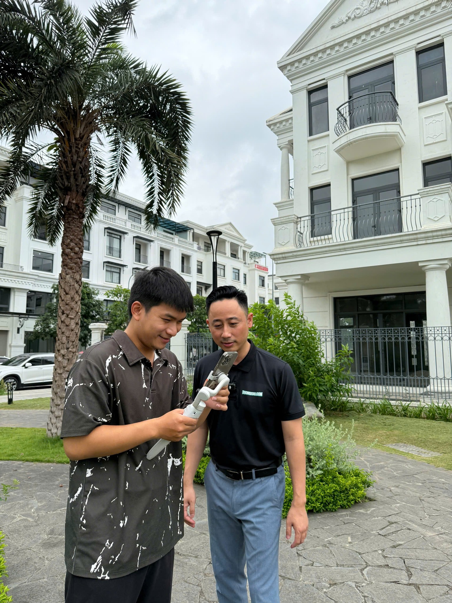Đăng Dương mặc áo polo đen đứng cùng một bạn nam cầm điện thoại gắn gimbal, cả hai cùng cúi xem lại đoạn vừa quay, phía sau là dãy biệt thự trắng và hàng cây cọ