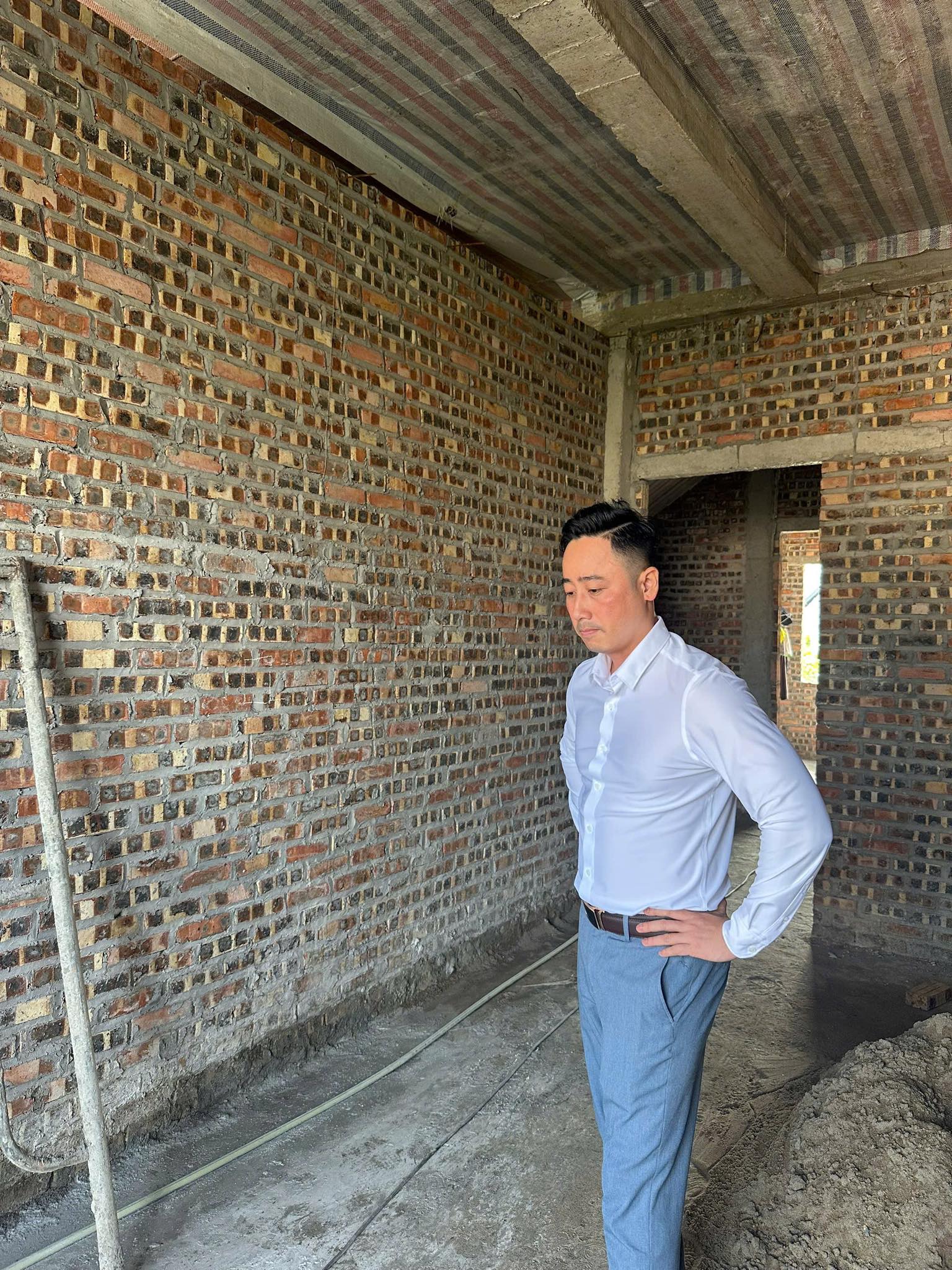 Đăng Dương mặc áo sơ mi trắng đứng bên trong một căn nhà mới xây thô, tường gạch chưa trát