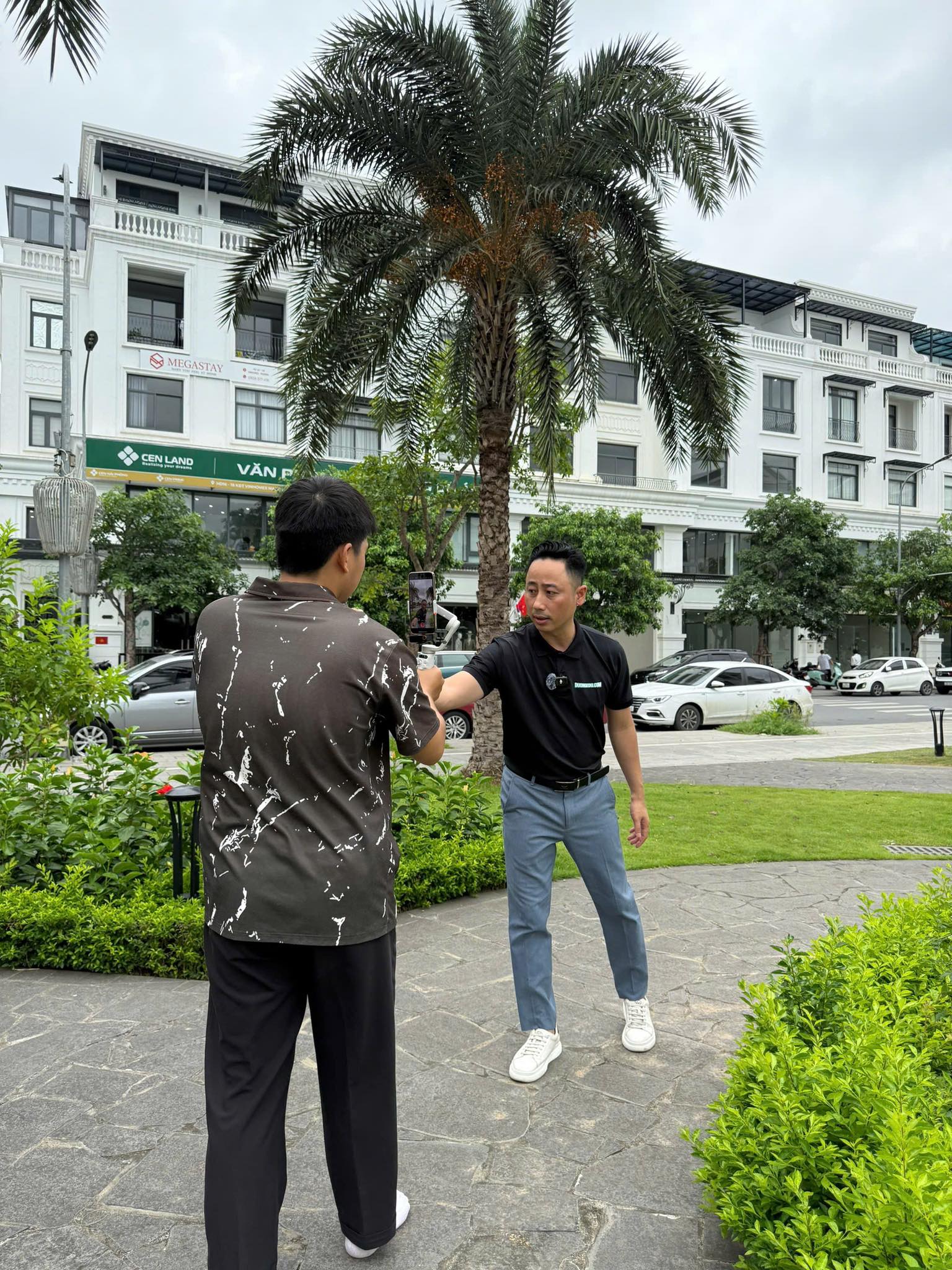 Đăng Dương mặc áo polo đen bắt tay một người đàn ông trên lối đi trong khu đô thị, phía sau là dãy nhà và văn phòng giao dịch
