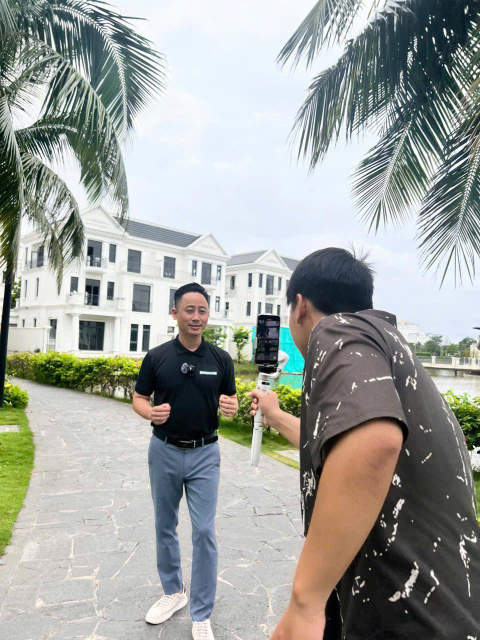 Đăng Dương mặc áo polo đen giơ hai ngón tay cái trước ống kính, một người cầm điện thoại gắn trên tay cầm chống rung quay lại, phía sau là dãy biệt thự