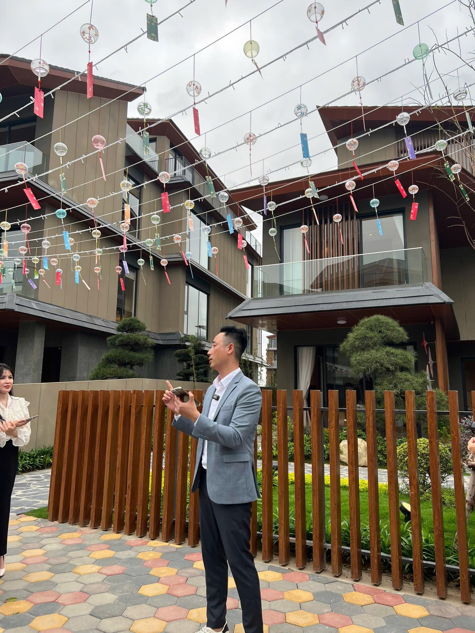 Đăng Dương mặc áo vest xám gài micro đứng nói và chỉ tay trước dãy biệt thự kiểu Nhật có giàn chuông gió, một người phụ nữ cầm điện thoại đứng nghe bên trái