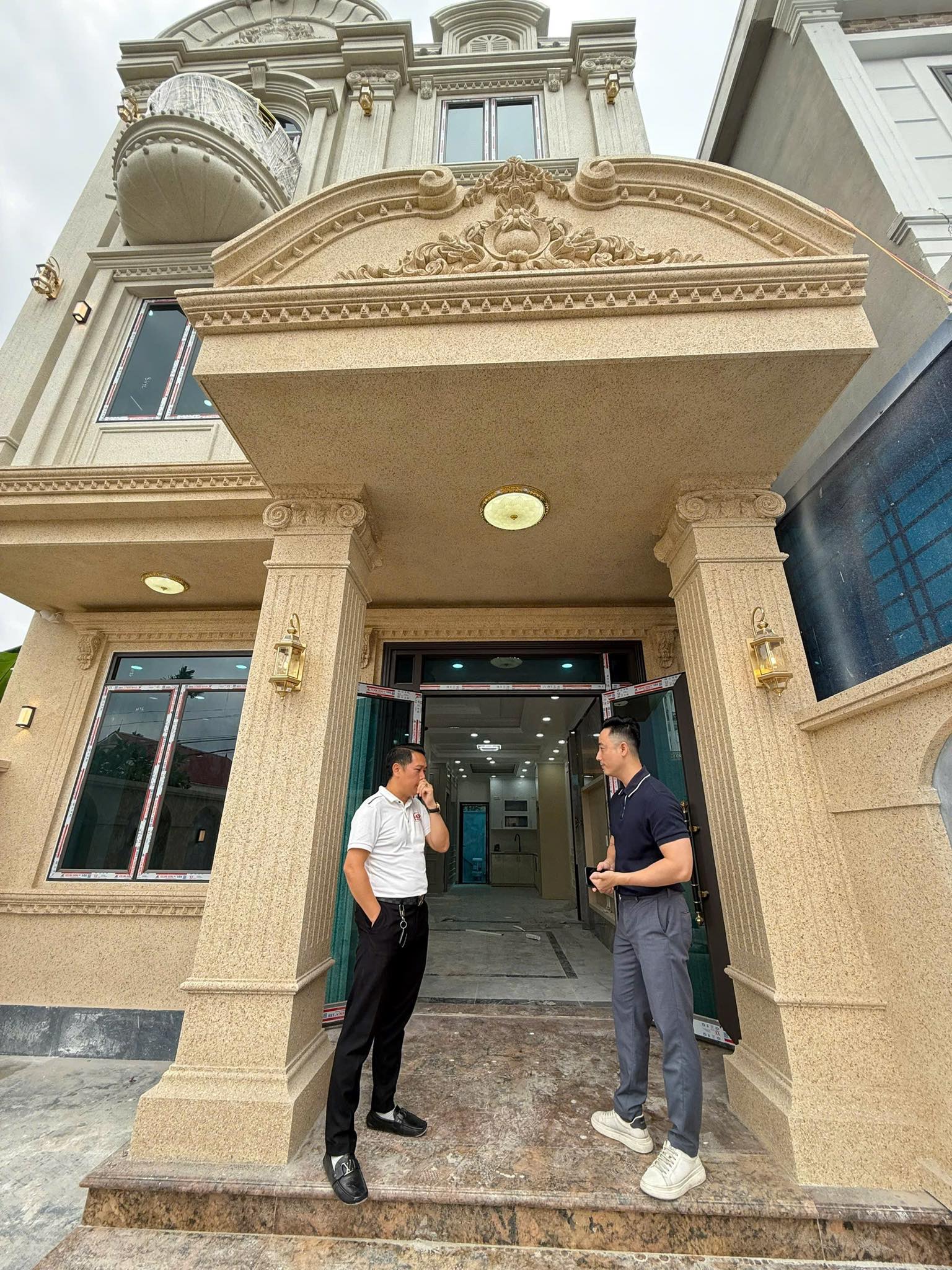 Đăng Dương mặc áo polo đen đứng trao đổi với một người đàn ông áo polo trắng trước sảnh một căn biệt thự tân cổ điển đang hoàn thiện