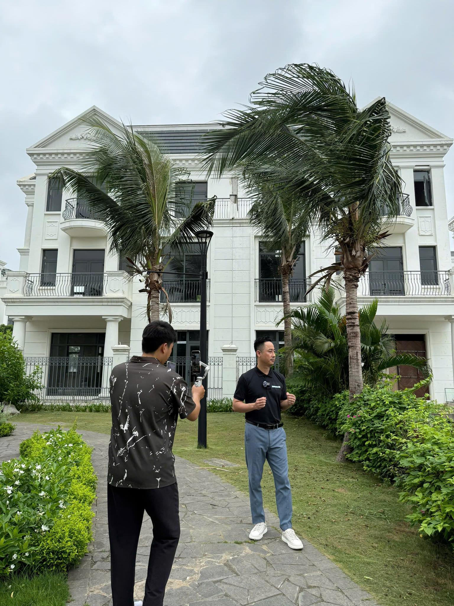 Đăng Dương mặc áo polo đen đứng nói trước ống kính khi một người trẻ cầm điện thoại gắn tay cầm chống rung quay, phía sau là biệt thự trắng ba tầng và hàng dừa