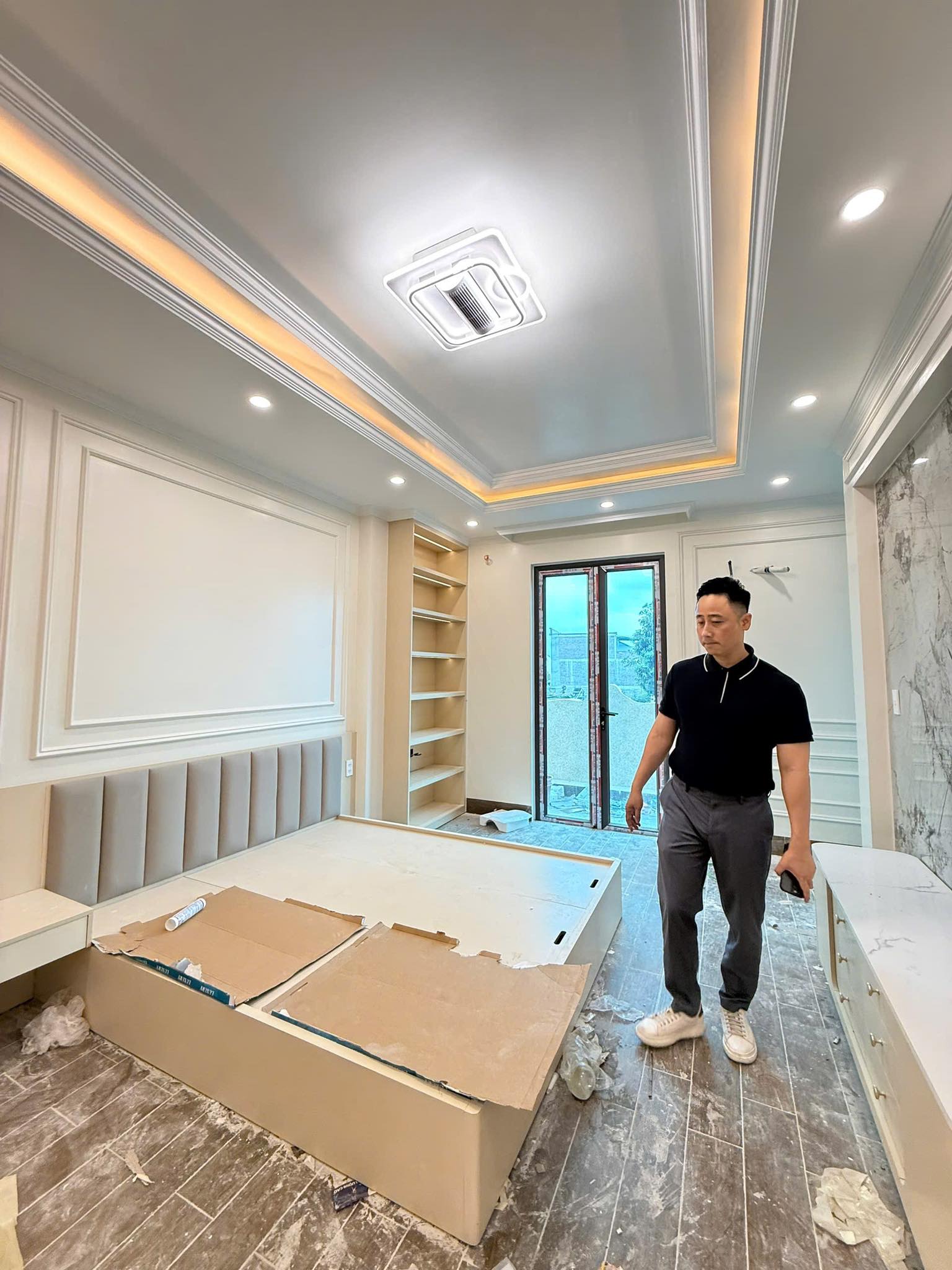 Đăng Dương mặc áo polo đen cầm điện thoại đi xem một phòng ngủ biệt thự đang hoàn thiện nội thất với trần phào chỉ và đèn hắt