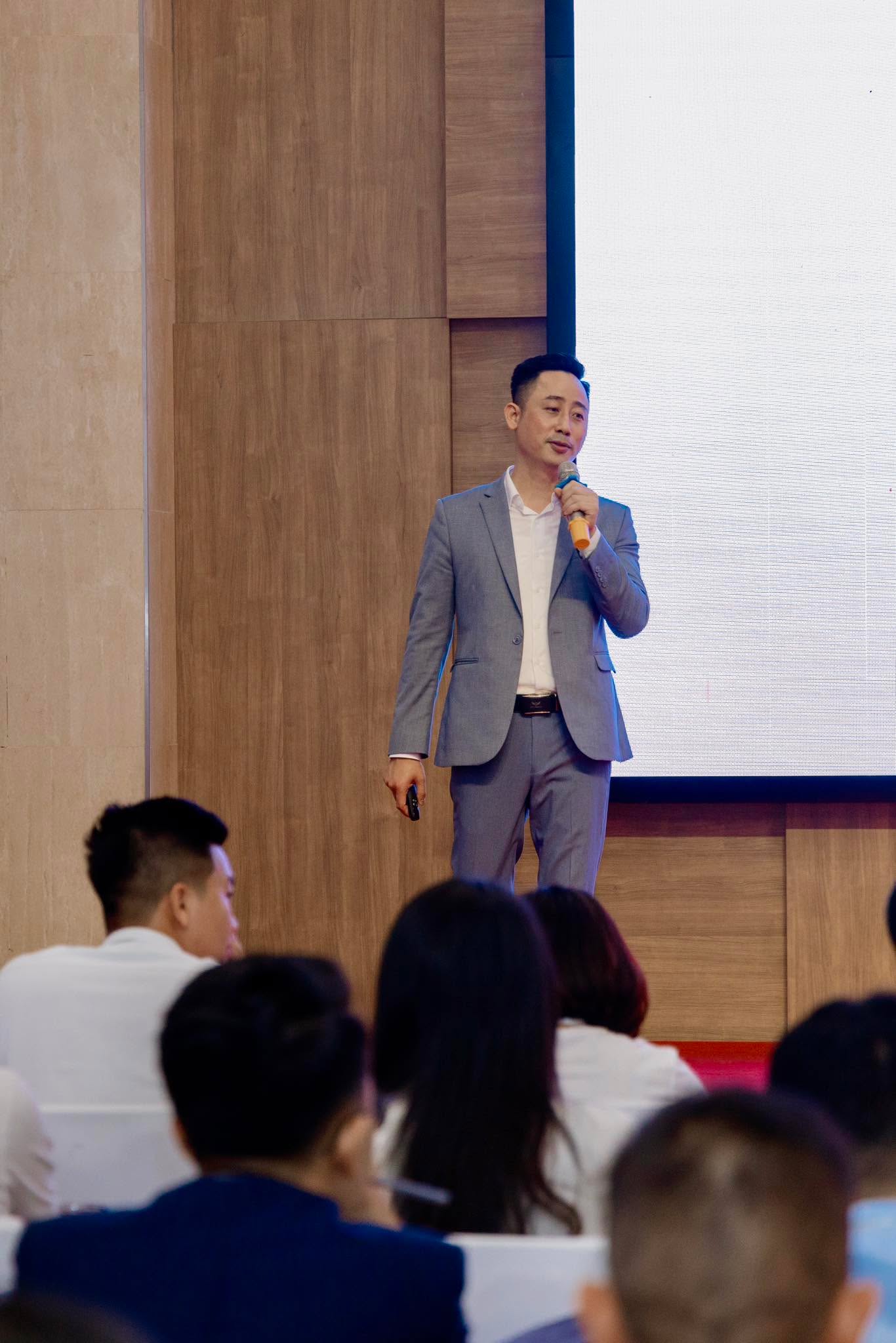 Đăng Dương mặc vest xám cầm micro đứng thuyết trình trên sân khấu hội trường, phía dưới là hàng ghế khán giả và bên phải là màn chiếu lớn