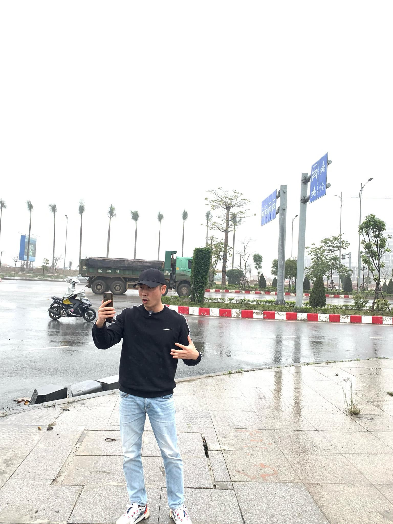 Đăng Dương đội mũ lưỡi trai đen mặc áo nỉ đen cầm điện thoại tự quay trên vỉa hè một đại lộ mới, phía sau là xe tải ben và hàng cau dọc dải phân cách