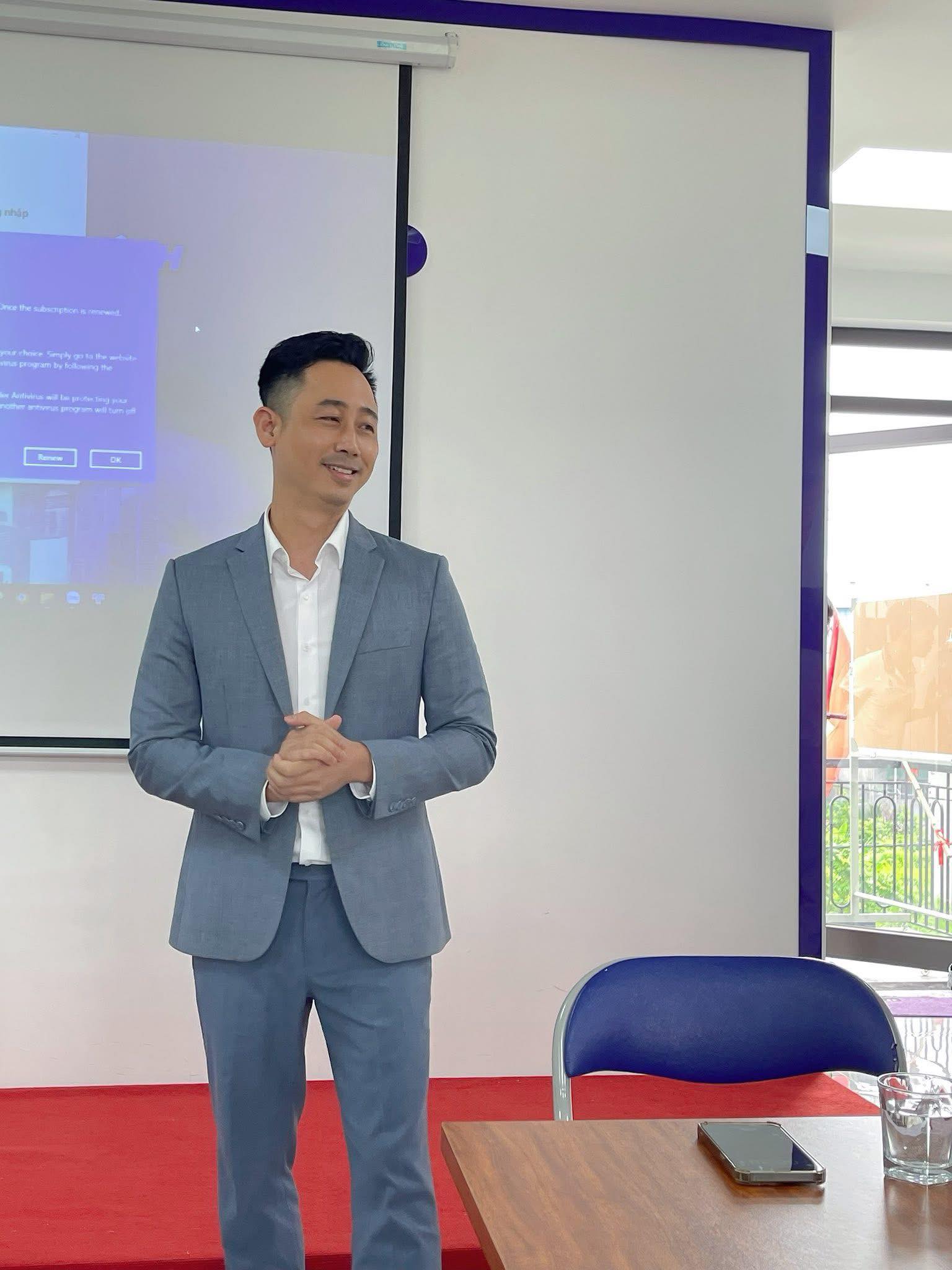 Đăng Dương mặc vest xám đứng cười trước màn chiếu trong một phòng họp nhỏ, bên cạnh là bàn gỗ và ghế xanh