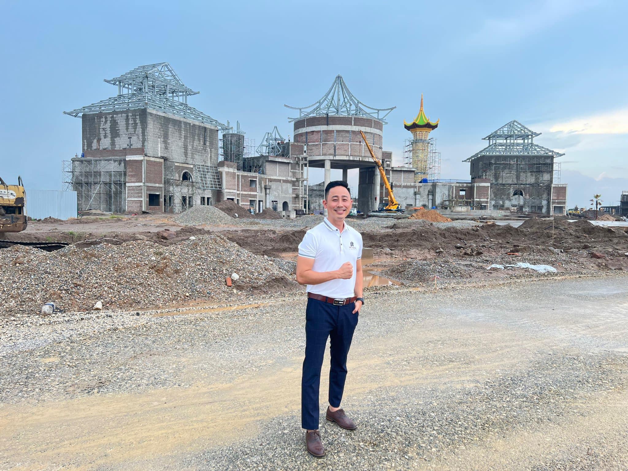 Đăng Dương mặc áo polo trắng đứng trên nền đất đá trước một công trình đang xây thô có cần cẩu