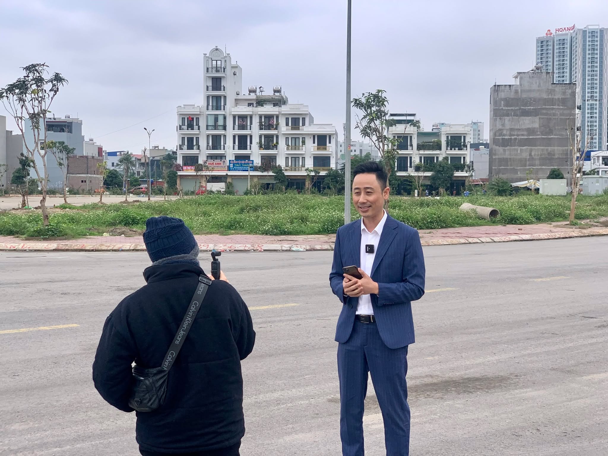 Đăng Dương mặc vest xanh than cầm điện thoại đứng nói bên lề đường trước một khu đất trống, một người đội mũ len quay lại bằng máy quay cầm tay