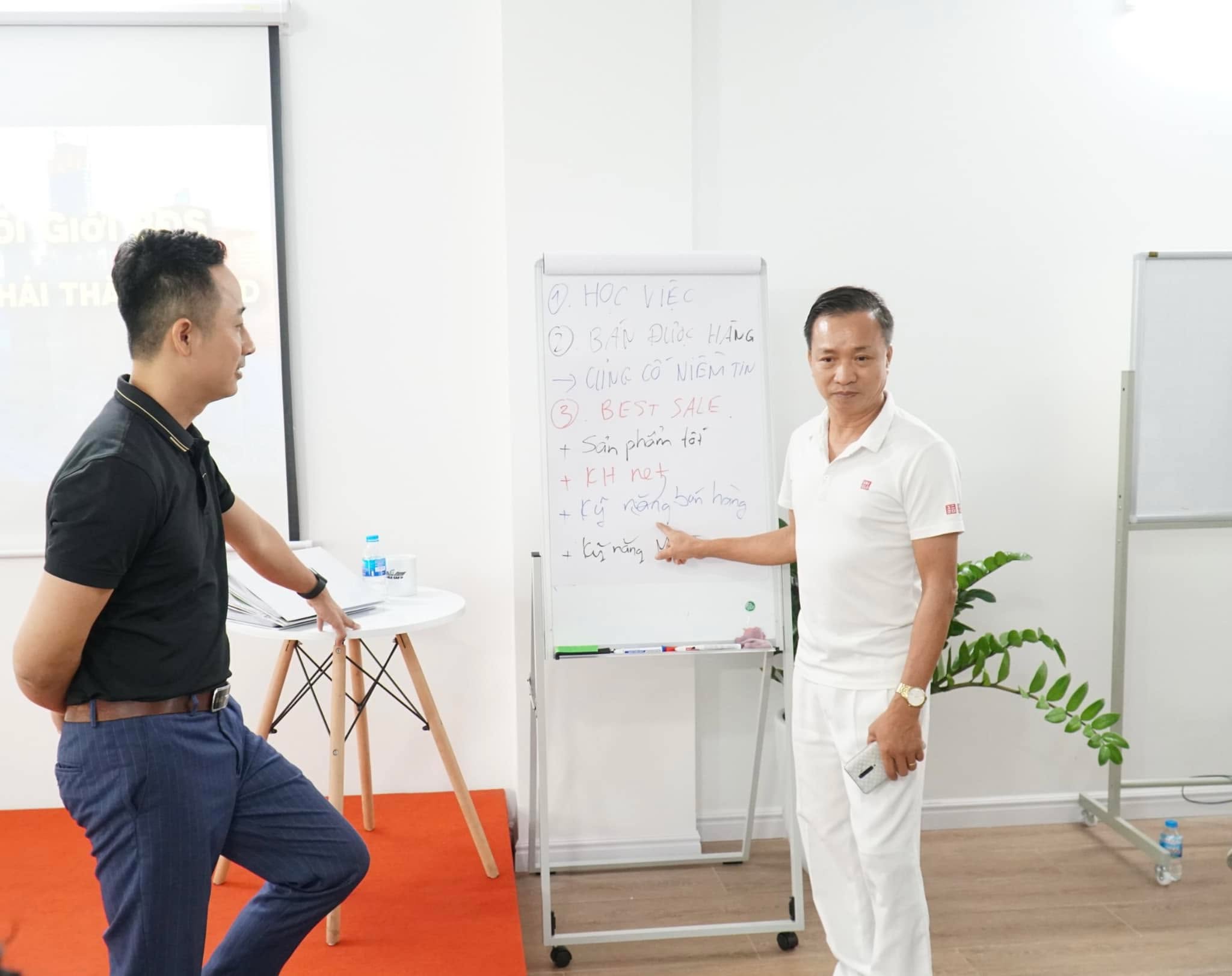 Đăng Dương mặc áo polo đen đứng nghe một người đàn ông áo trắng chỉ vào bảng lật ghi các bước học việc, bán được hàng và best sale