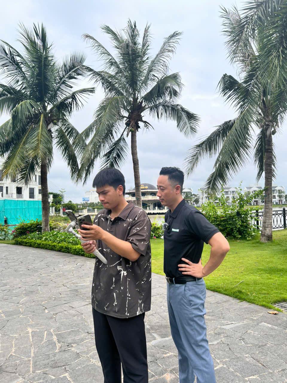 Đăng Dương mặc áo polo đen chống nạnh đứng xem một người trẻ cầm điện thoại gắn trên tay cầm chống rung, phía sau là hàng dừa trong khu đô thị