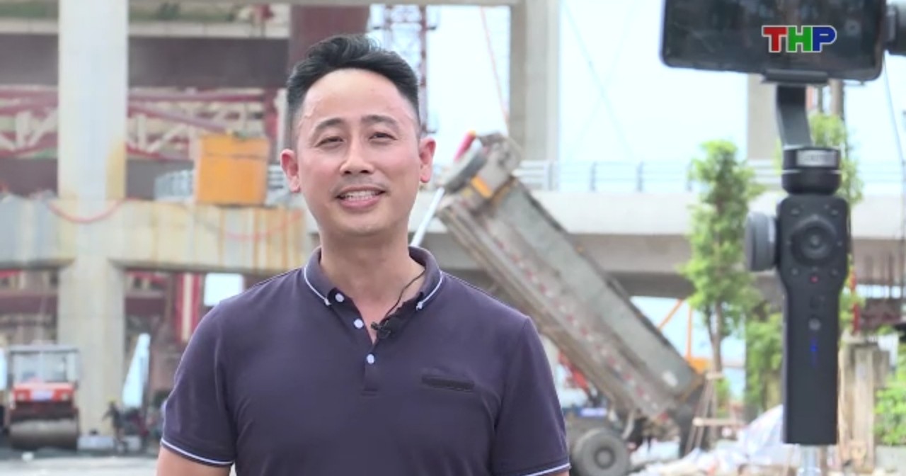Đăng Dương mặc áo polo tím than đứng nói trước chiếc điện thoại gắn trên gimbal, phía sau là công trường cầu đang thi công và một xe ben đang đổ đất