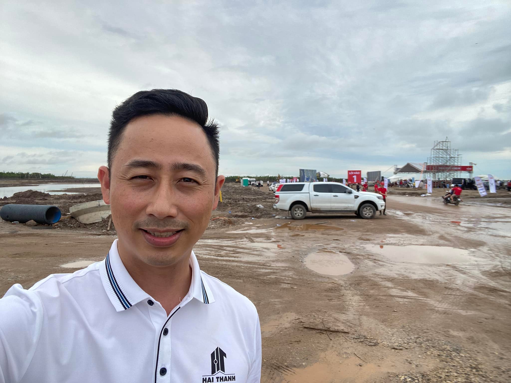 Đăng Dương mặc áo polo trắng tự chụp ảnh tại một khu đất dự án vừa san nền còn lầy bùn, phía sau là xe bán tải trắng và khung giàn sân khấu sự kiện