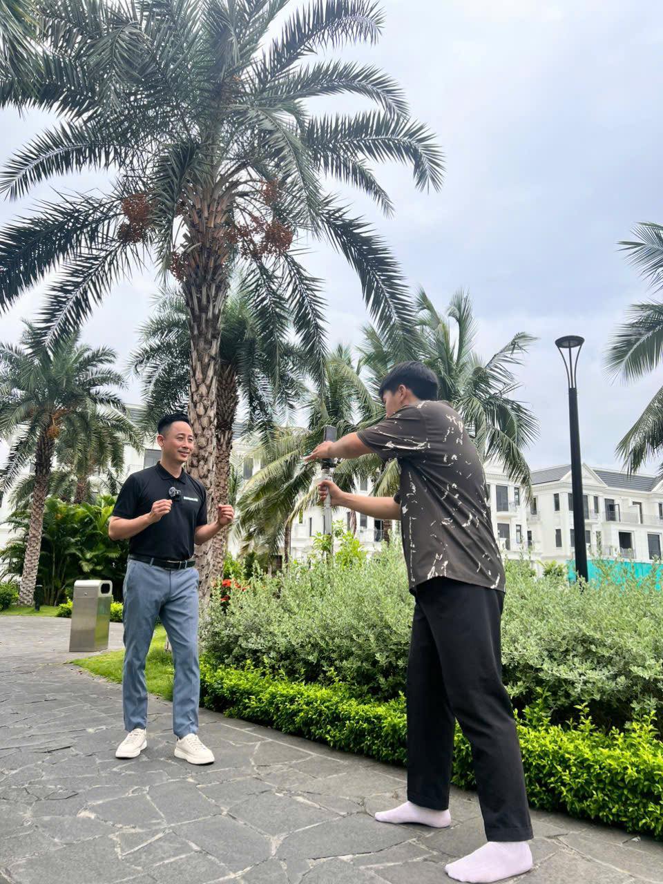 Đăng Dương mặc áo polo đen đứng nói trước một người đang cầm điện thoại gắn gimbal quay lại, dưới hàng cây cọ trong khu đô thị