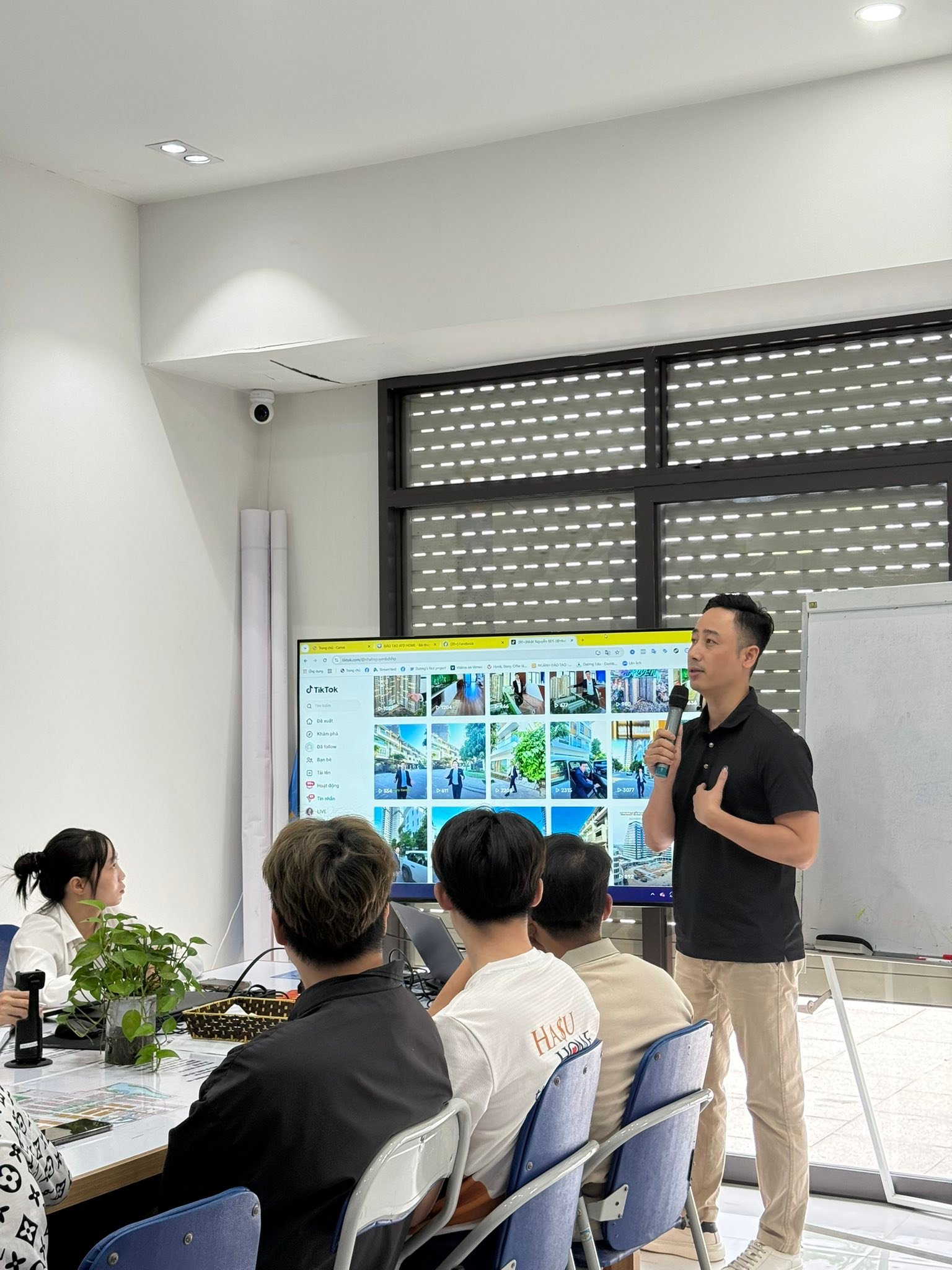 Đăng Dương mặc áo polo đen cầm micro đứng nói bên cạnh màn hình lớn đang mở lưới video TikTok, phía trước là nhóm học viên ngồi nghe