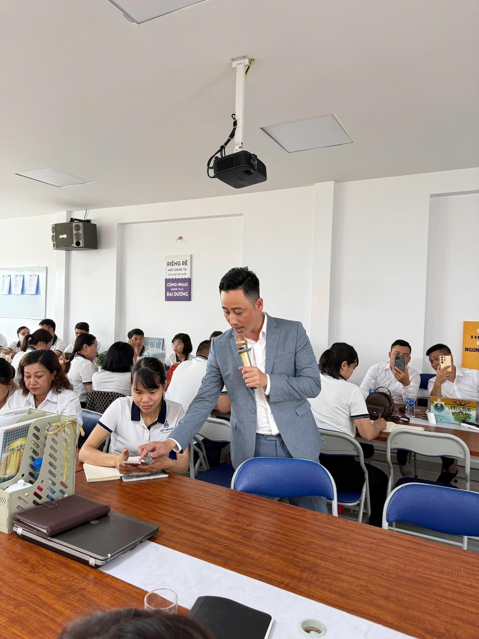 Đăng Dương mặc vest xám cầm micro cúi xuống trao đổi riêng với một học viên đang ghi chép, trong lớp học đông người mặc áo đồng phục trắng