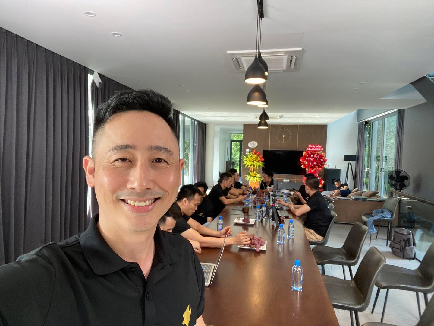 Đăng Dương mặc áo polo đen tự chụp ảnh trong phòng họp có bàn dài, phía sau là nhóm đồng nghiệp áo đen ngồi làm việc với laptop và một điện thoại gắn trên chân máy
