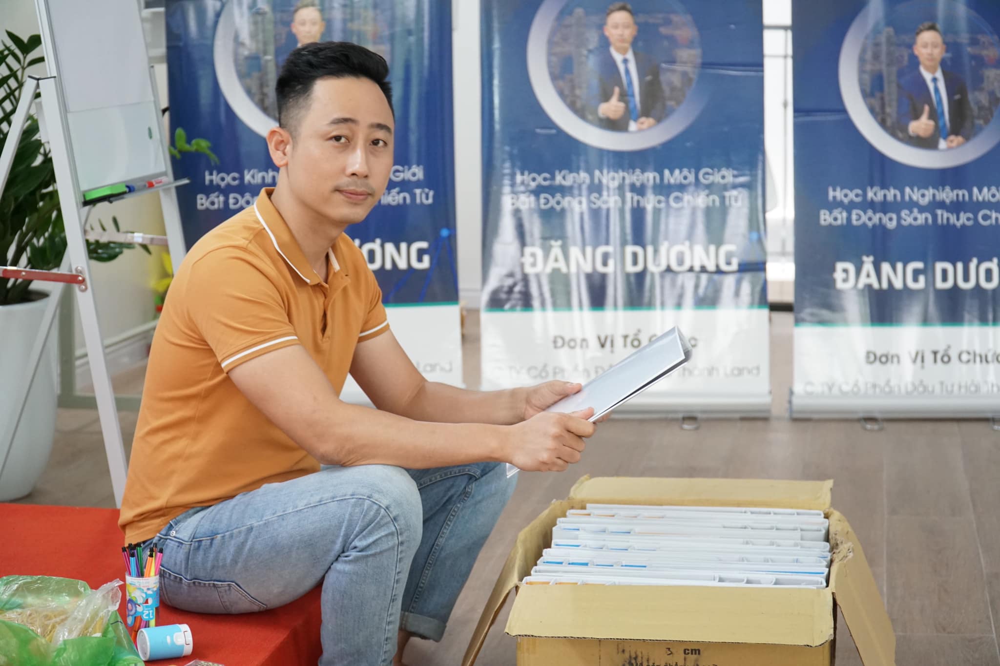 Đăng Dương mặc áo polo cam ngồi cạnh thùng tài liệu đang cầm một tập giấy, phía sau là ba standee in dòng chữ Học Kinh Nghiệm Môi Giới Bất Động Sản Thực Chiến Từ Đăng Dương