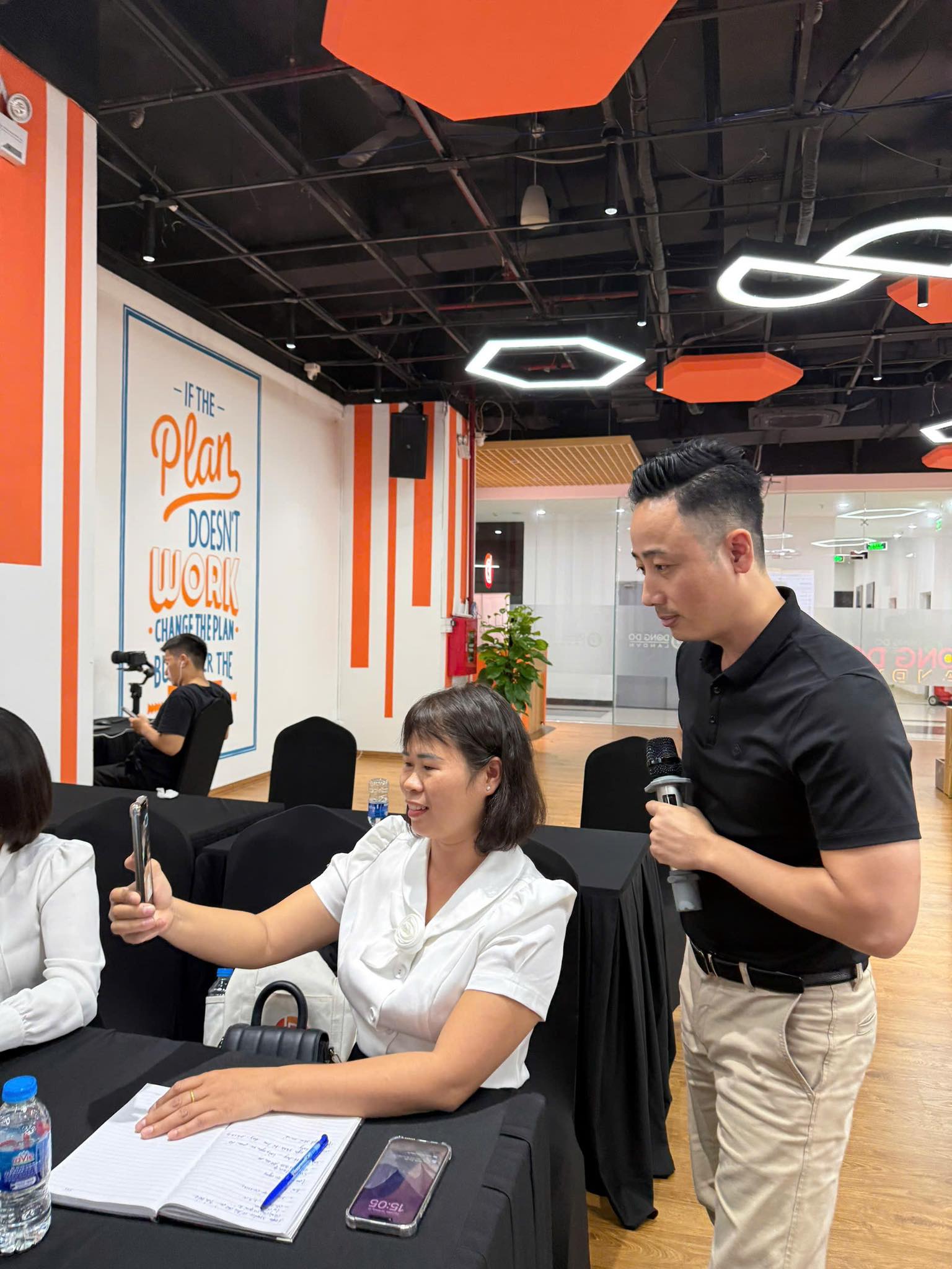 Đăng Dương mặc áo polo đen cầm micro đứng cạnh một học viên nữ đang giơ điện thoại lên quay trong phòng đào tạo tường cam