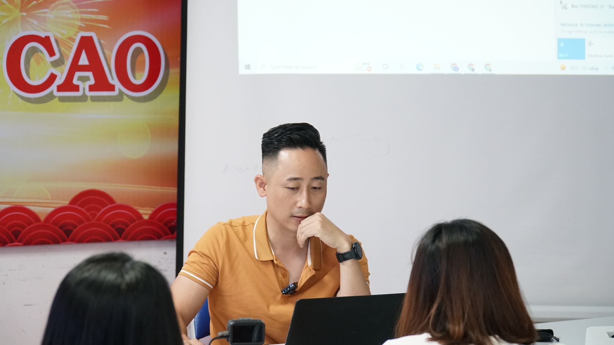 Đăng Dương mặc áo polo cam ngồi ở bàn, tay chống cằm suy nghĩ, phía trước là hai học viên ngồi trước màn hình máy tính