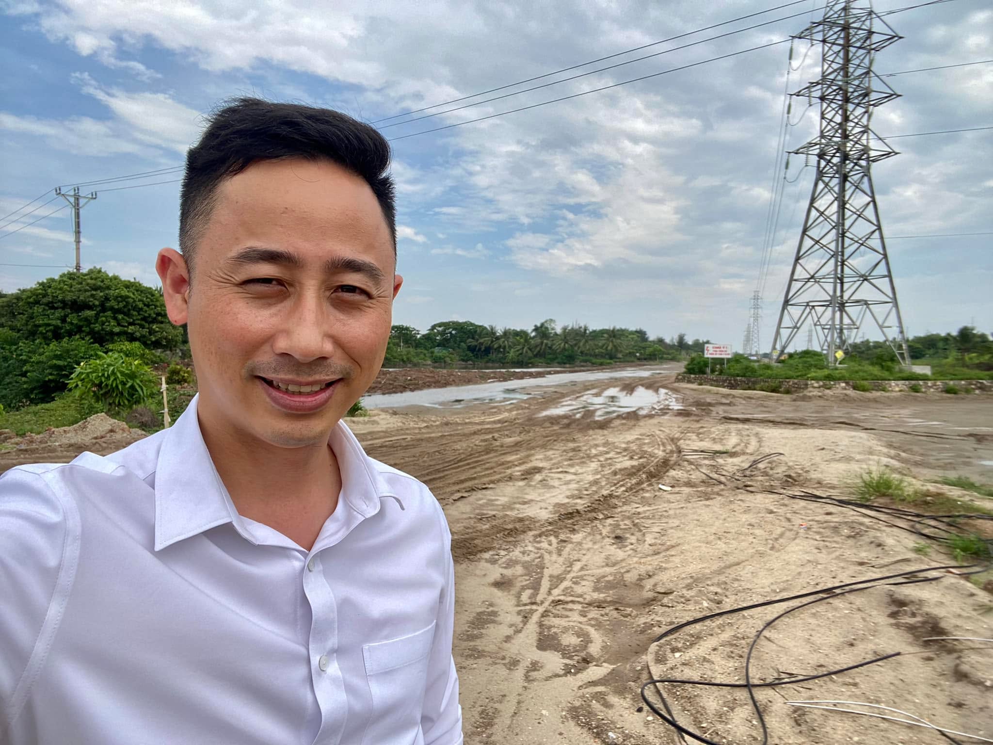 Đăng Dương mặc áo sơ mi trắng tự chụp ảnh trước một con đường đất đang san lấp, phía xa là cột điện cao thế
