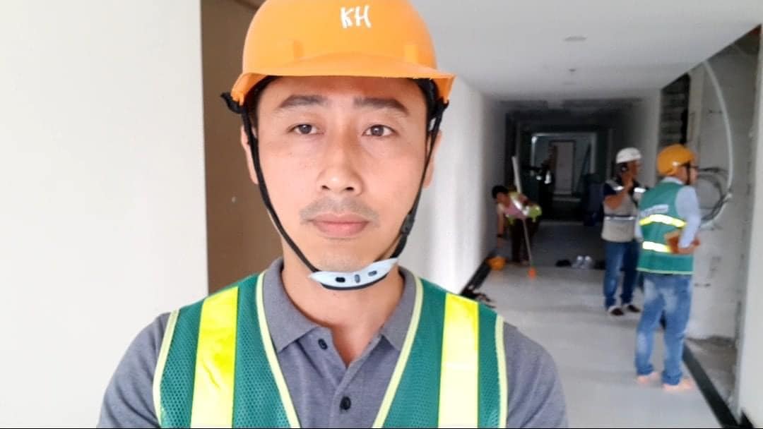 Đăng Dương đội mũ bảo hộ màu cam và mặc áo phản quang đứng trong một toà nhà đang hoàn thiện, phía sau là những công nhân đang làm việc