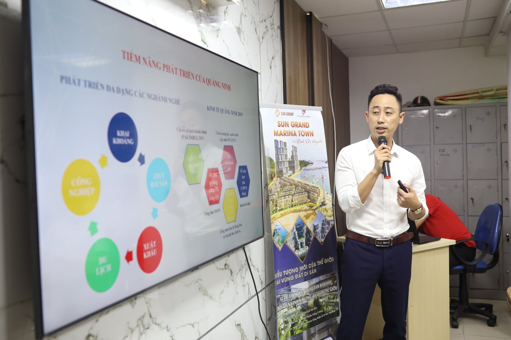 Đăng Dương mặc áo sơ mi trắng cầm micro thuyết trình cạnh màn hình lớn đang chiếu slide tiềm năng phát triển của Quảng Ninh, bên cạnh là standee giới thiệu một dự án