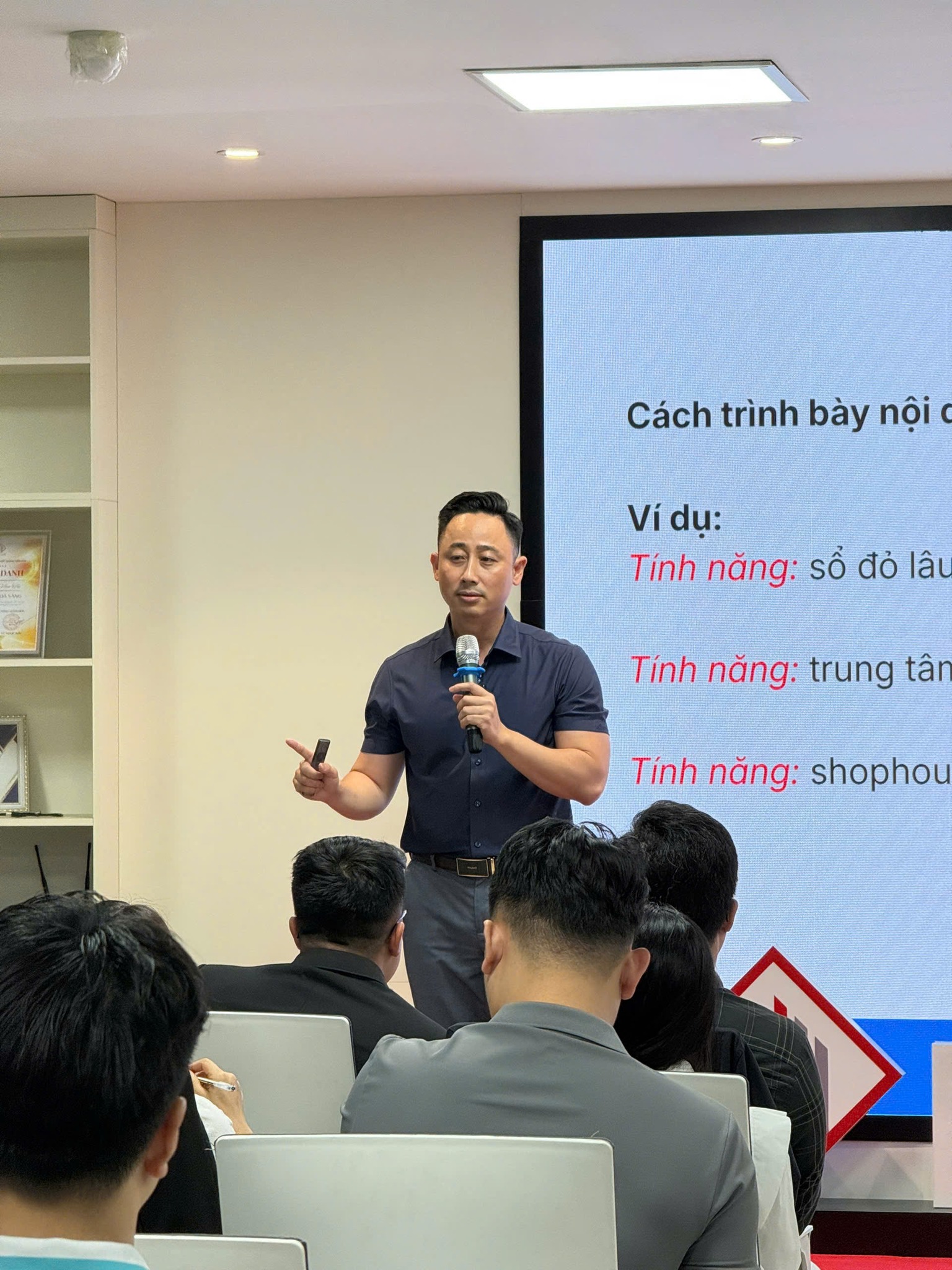 Đăng Dương mặc áo polo tối màu cầm micro đứng giảng trước màn chiếu nội dung về cách trình bày tính năng bất động sản, phía trước là học viên ngồi nghe