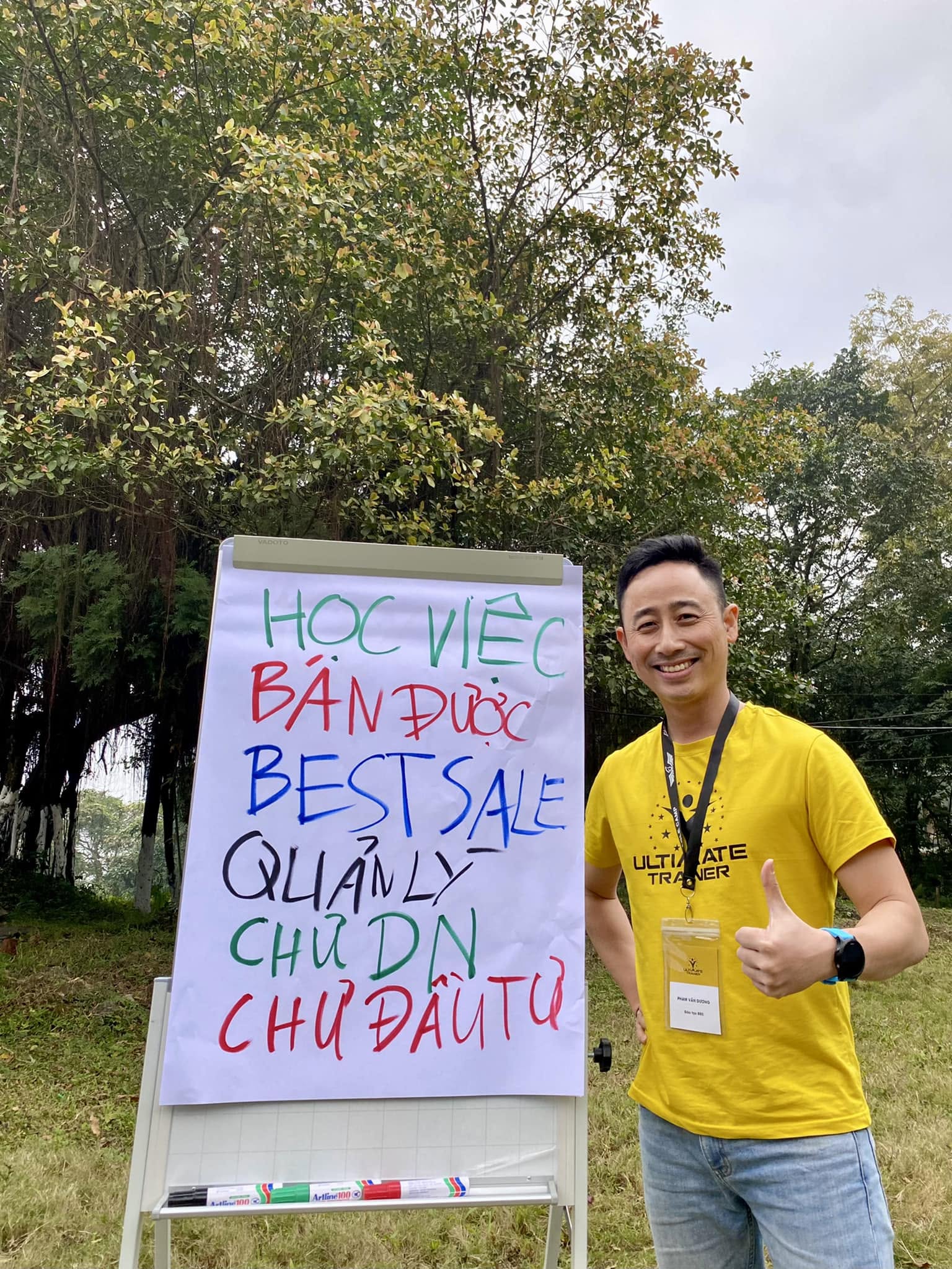 Đăng Dương đứng cạnh bảng flipchart ghi các cấp bậc nghề môi giới bất động sản trong một buổi đào tạo ngoài trời