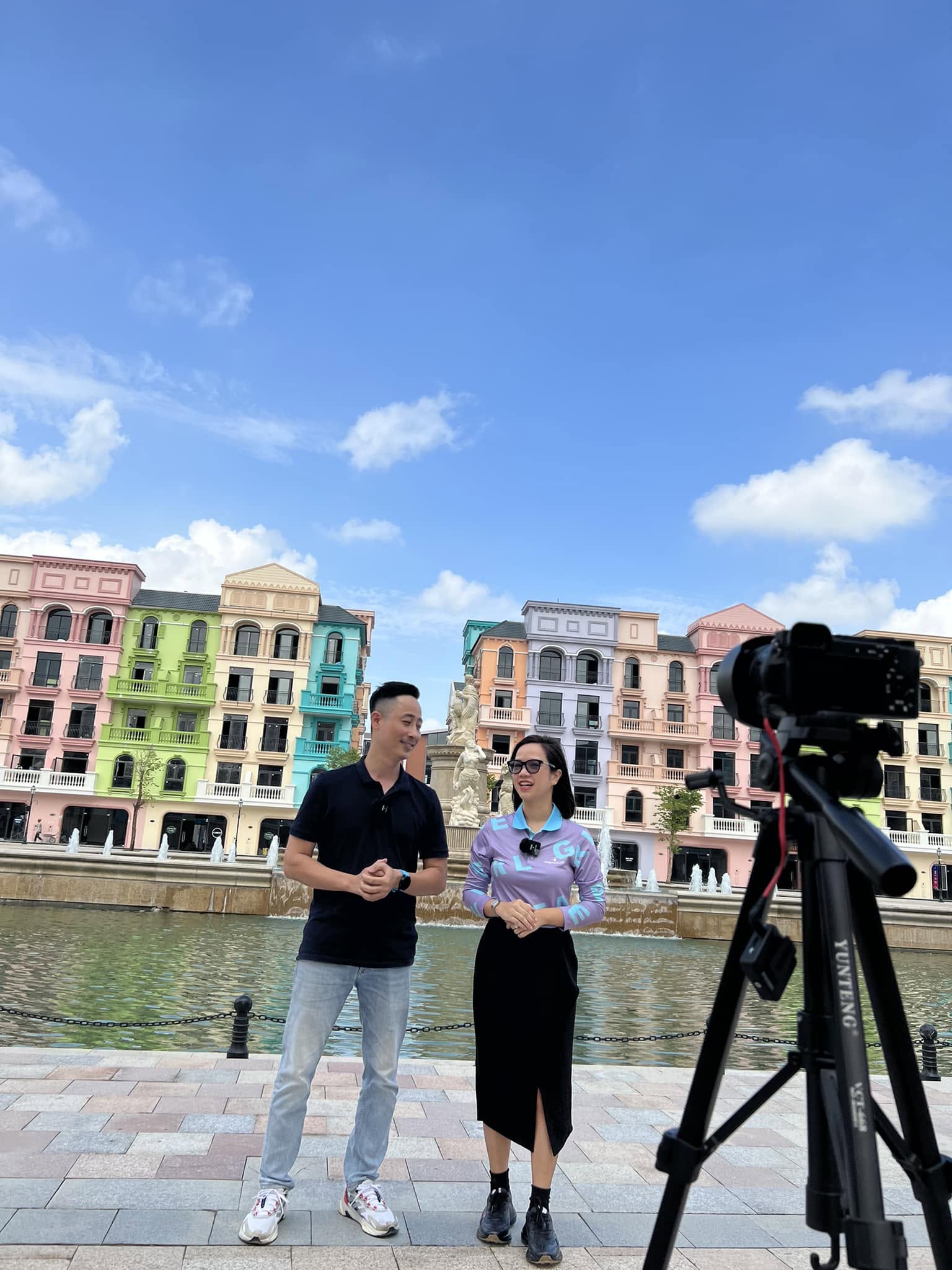 Đăng Dương và một đồng nghiệp giới thiệu trước máy quay tại một dự án nhà phố nhiều màu sắc, phía sau là hồ nước có đài phun nước