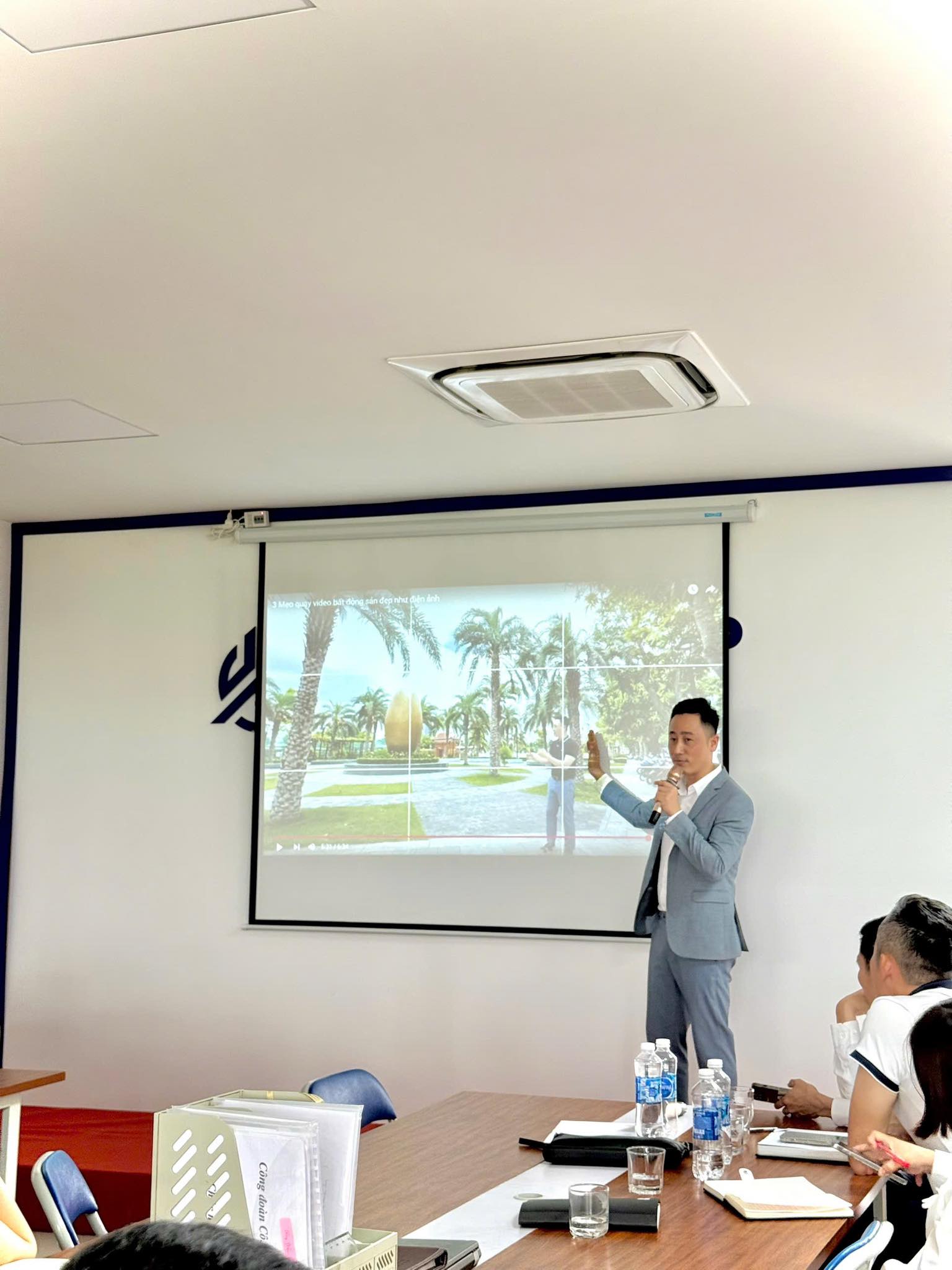 Đăng Dương mặc vest xám cầm micro đứng giảng trong phòng họp, phía sau là màn chiếu đang phát một video bất động sản