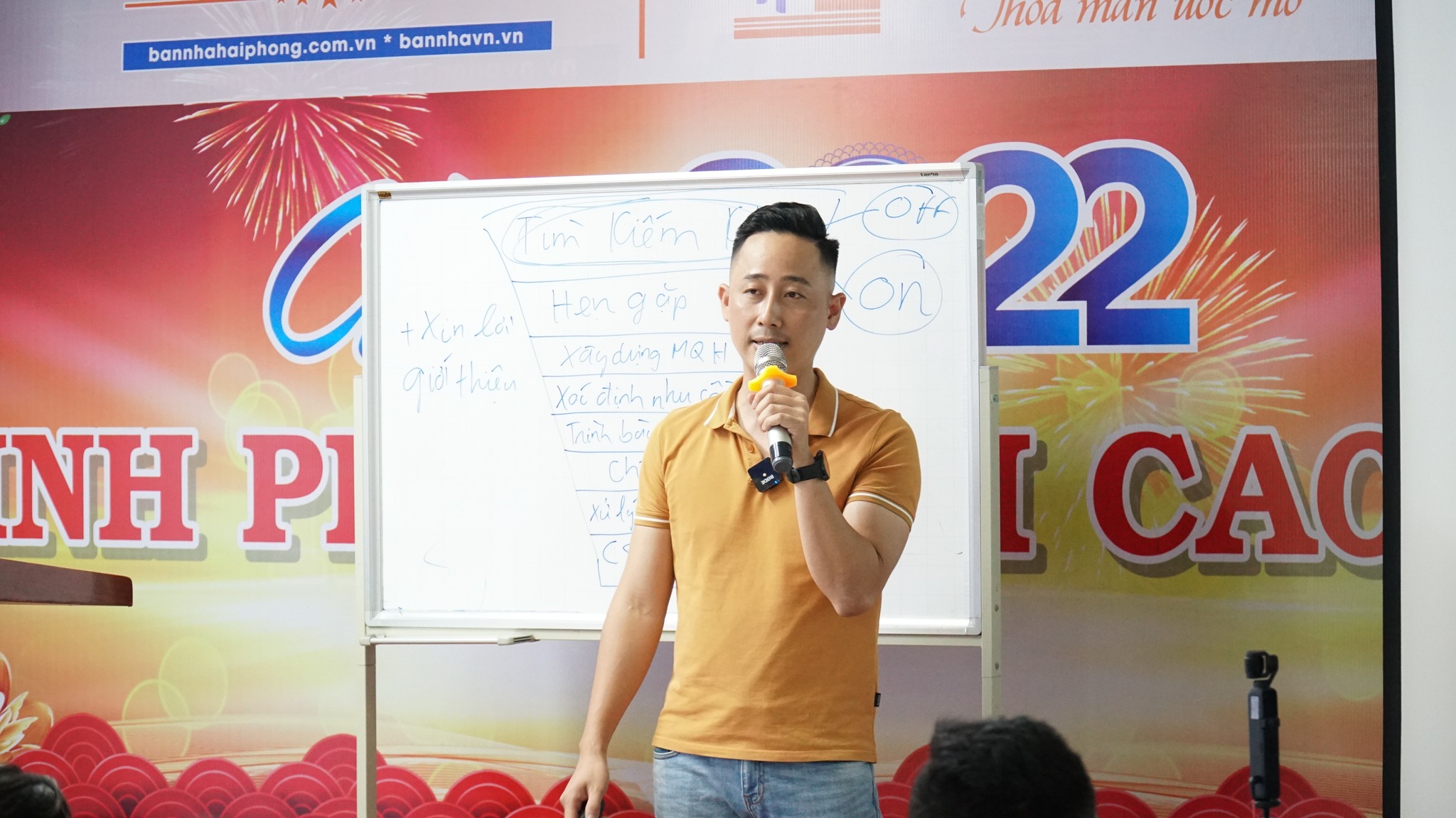 Đăng Dương mặc áo polo vàng cầm micro đứng nói cạnh một tấm bảng trắng vẽ sơ đồ phễu quy trình bán hàng, phía sau là phông sân khấu