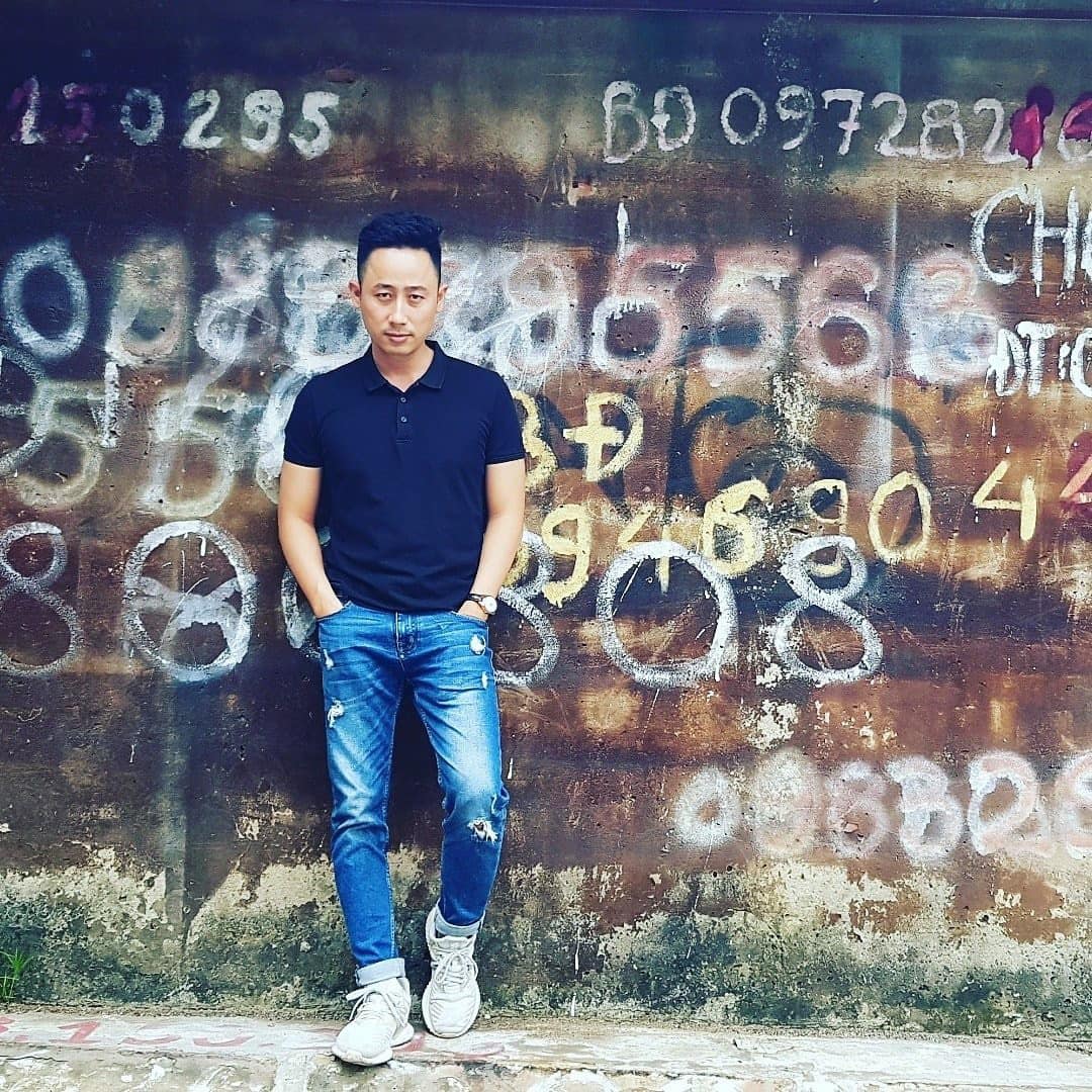 Đăng Dương mặc áo polo đen và quần jeans đứng tựa một bức tường cũ phủ đầy số điện thoại rao bán đất viết bằng sơn