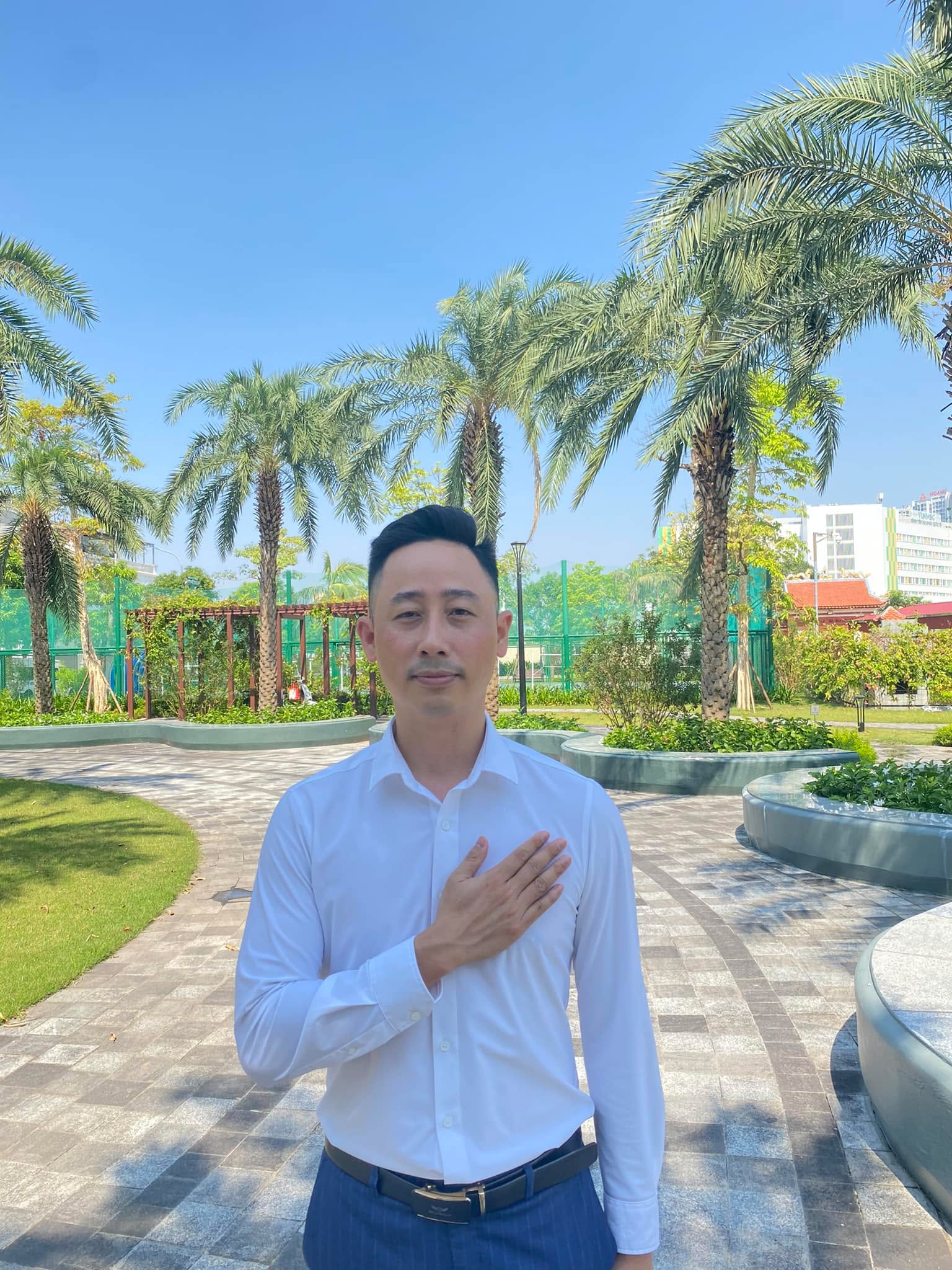 Đăng Dương mặc áo sơ mi trắng đặt tay lên ngực đứng trên lối đi lát gạch giữa công viên nội khu một khu đô thị có hàng cây cọ