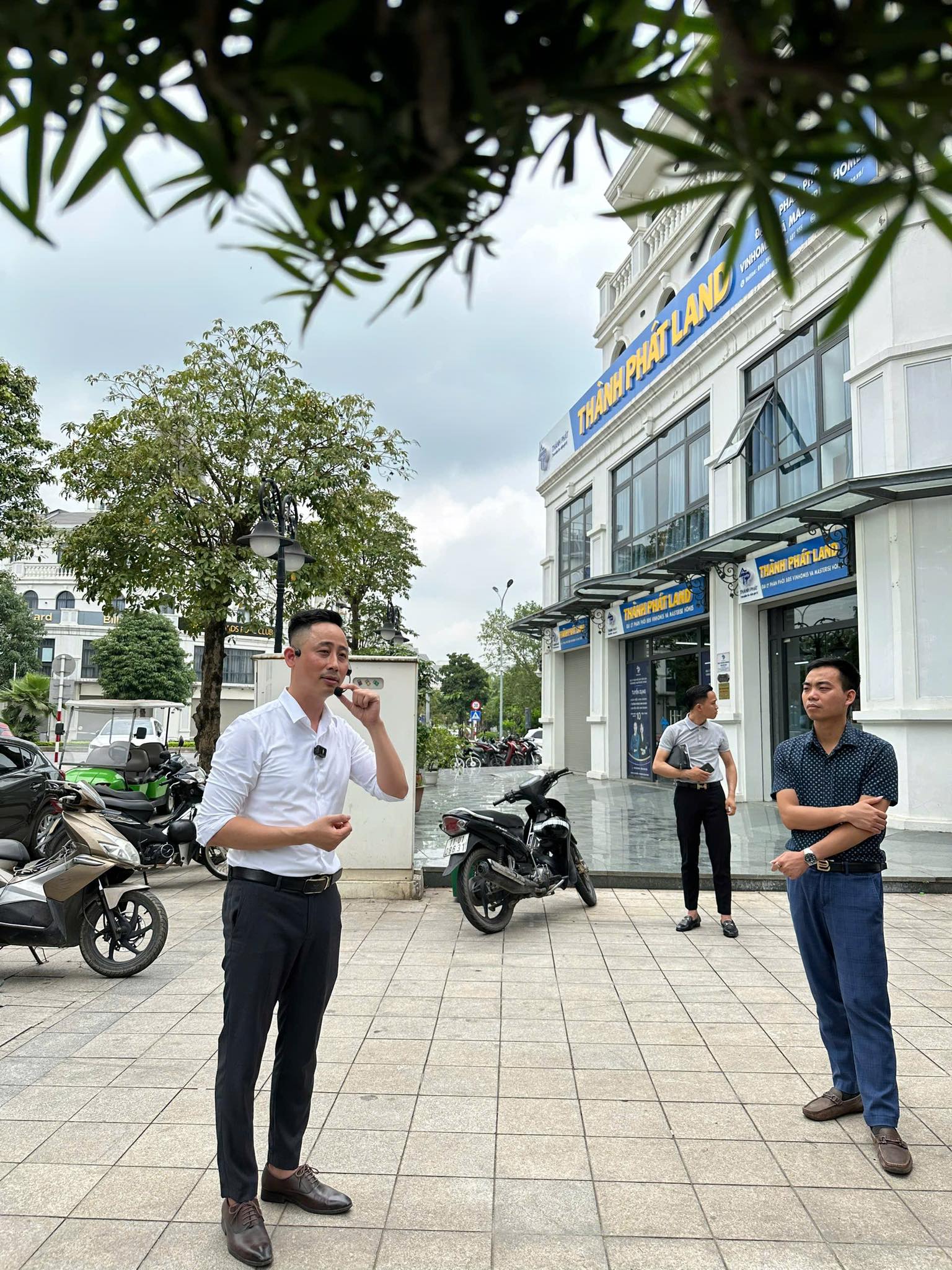Đăng Dương mặc áo sơ mi trắng đeo micro cài áo đang quay video ngoài quảng trường trước một văn phòng giao dịch bất động sản, hai người đứng nghe bên cạnh